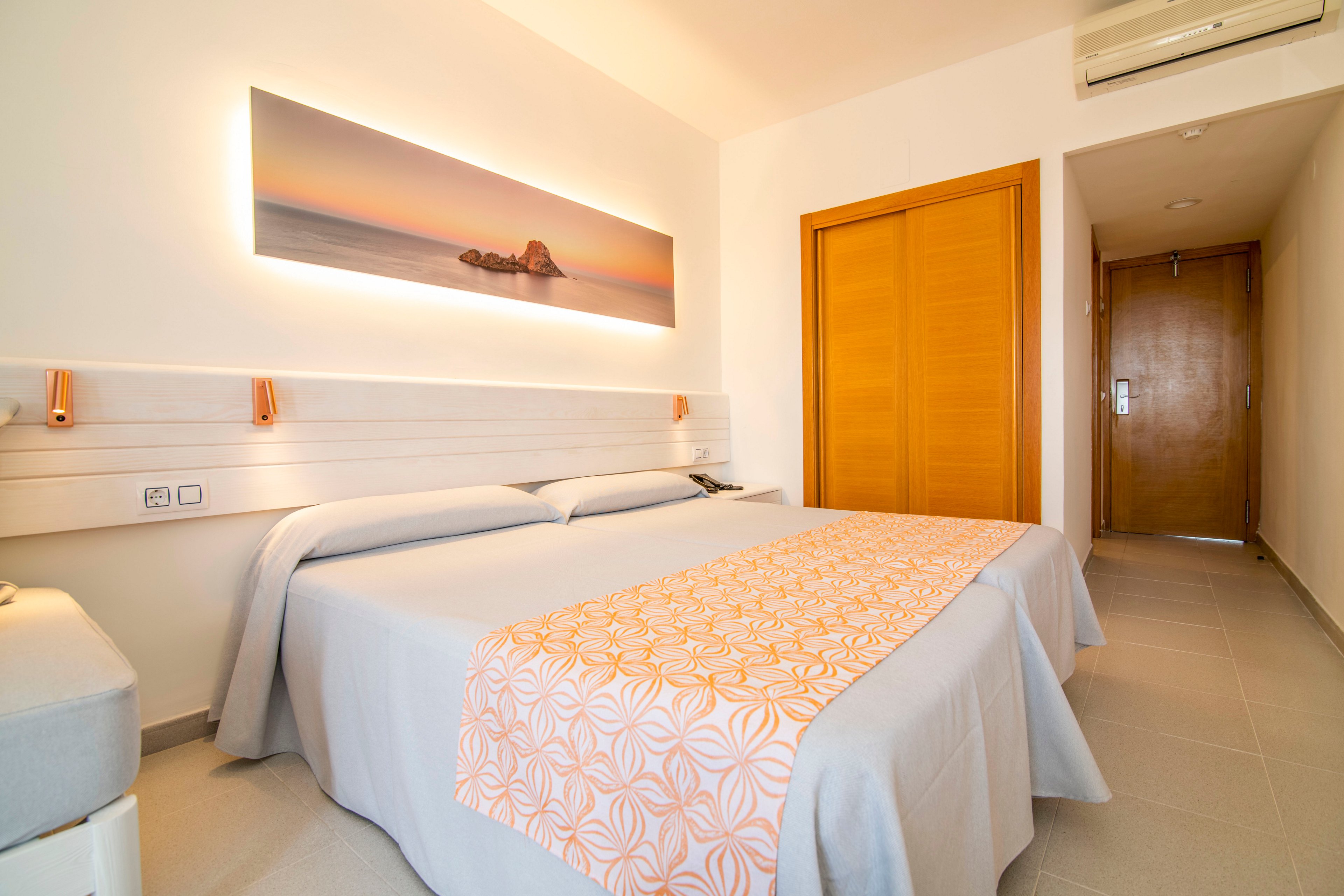 A clean, modern hotel room features a large double bed with light gray bedding and an orange patterned runner. Warm wooden elements, including a sliding closet door and entry door, contrast with the neutral walls and floor. Above the bed hangs a panoramic artwork of a serene sunset over the sea, adding a relaxing touch. Soft lighting and minimal decor create a calm, inviting atmosphere suitable for guests.