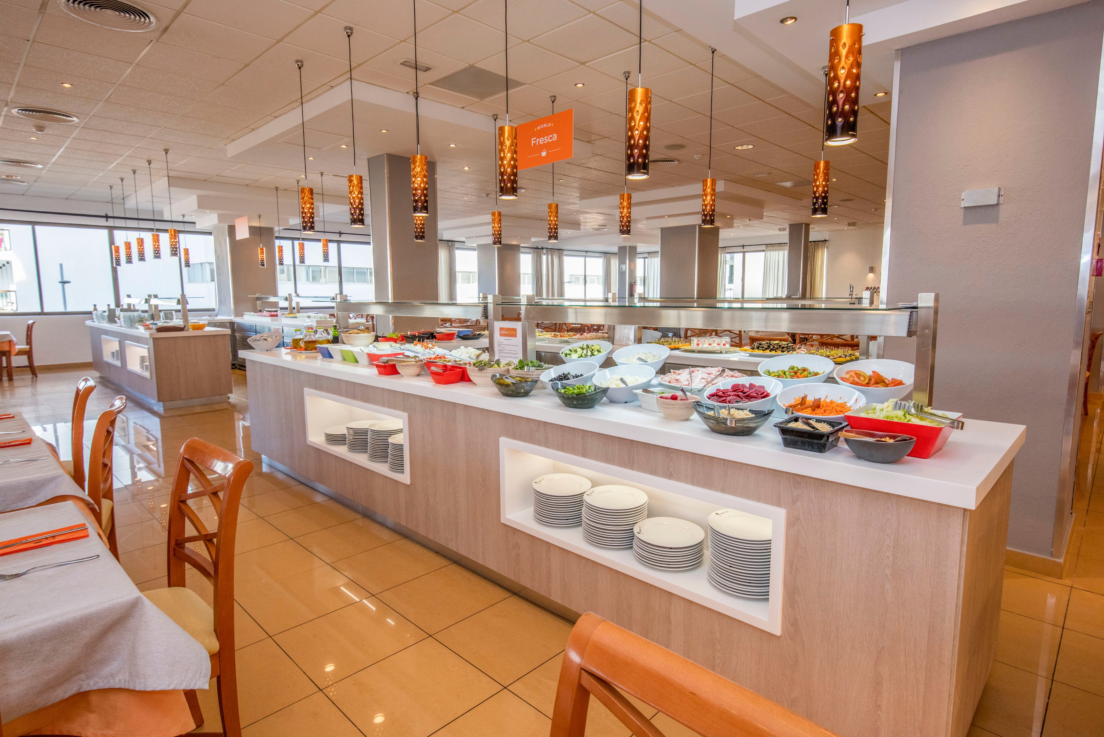 A spacious self-service buffet area displays a wide variety of salads and cold dishes in bowls and trays. The counter is neatly organized with stacks of plates and serving utensils ready for guests. Warm pendant lights hang from the ceiling, illuminating the modern dining room with large windows and tiled floors. Wooden chairs and tables with neatly arranged table settings create a clean, inviting restaurant atmosphere.