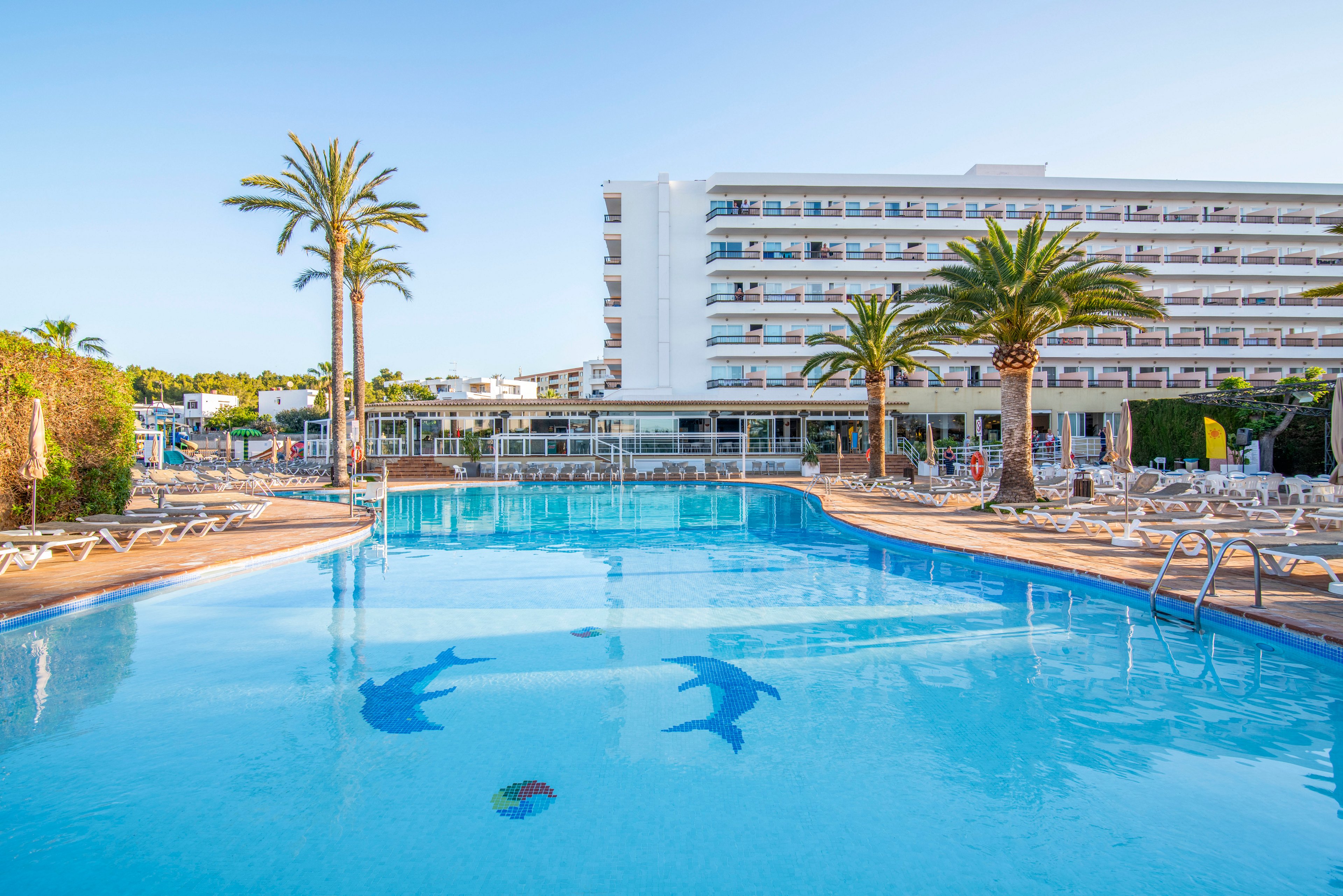 A large resort swimming pool dominates the foreground, reflecting the clear blue sky and surrounding palm trees. Two dolphin mosaics are visible on the pool floor, adding a playful decorative element. Sun loungers and parasols line the poolside area, suggesting a relaxing vacation setting. In the background, a multi-story hotel building with balconies overlooks the tranquil scene.