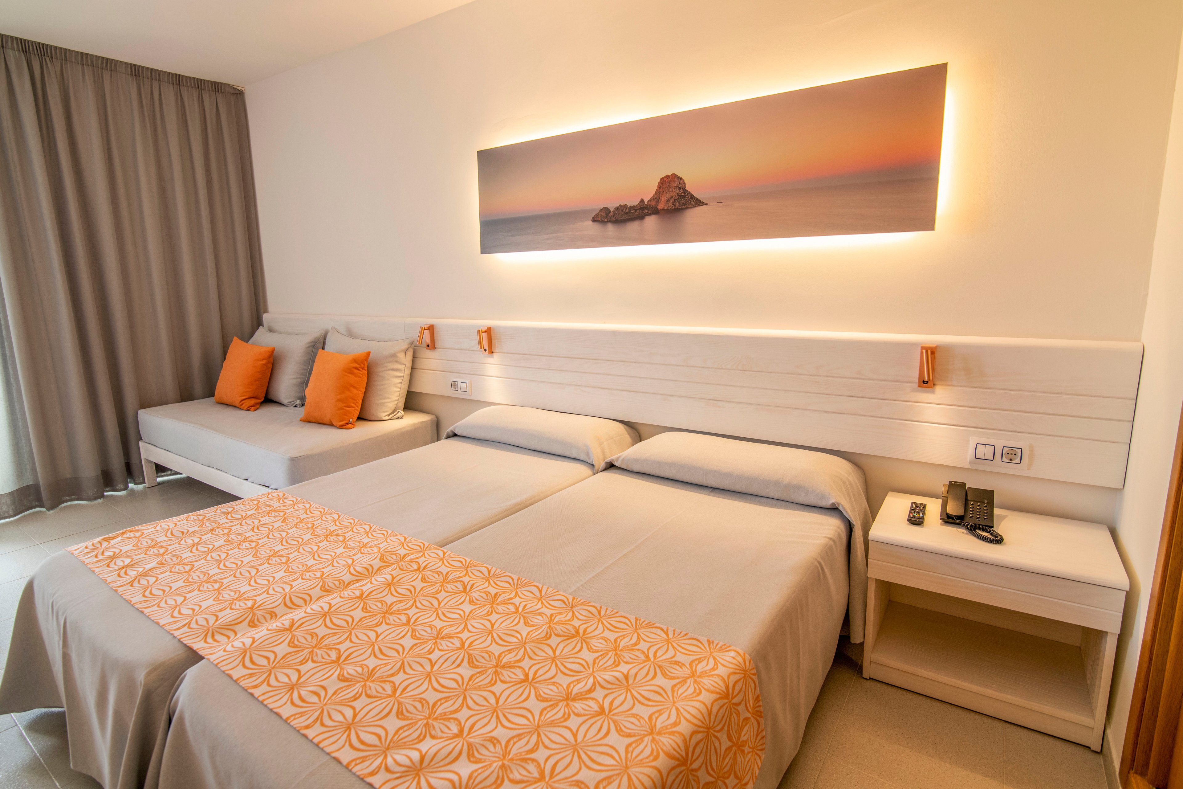 Bright contemporary hotel room featuring two neatly made twin beds with patterned orange runners. A sofa daybed with orange accent pillows sits beside the beds near floor-length curtains. Warm ambient lighting highlights a panoramic wall art of a serene seascape above the headboard. A bedside table holds a telephone and remote control, emphasizing a clean and organized guest space.