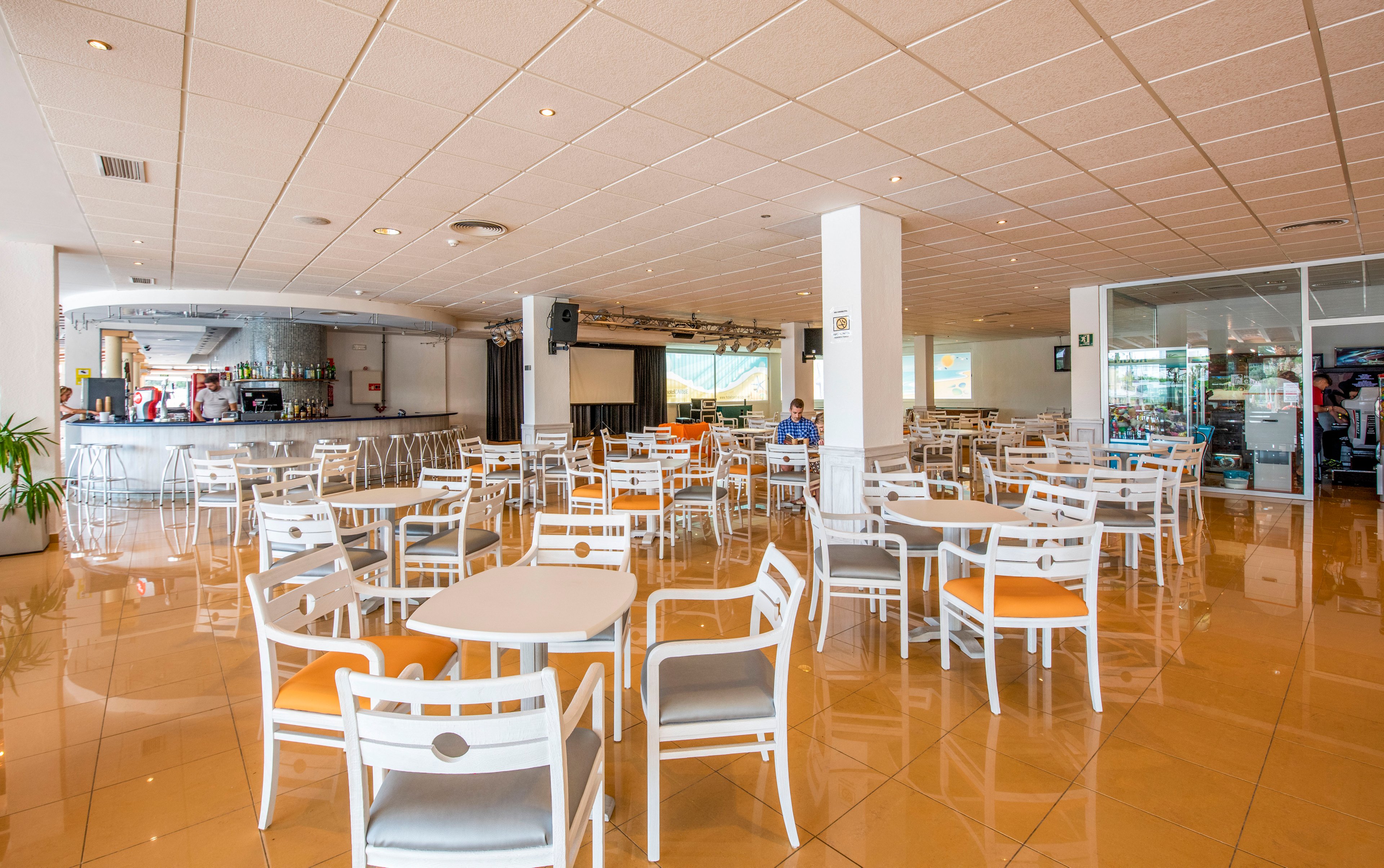 Spacious cafeteria interior with white tables and chairs featuring orange seat cushions. The room has a glossy tiled floor and a low, grid-pattern ceiling with recessed lighting. A bar counter with stools and drink displays is visible in the background. Large windows and glass partitions bring in natural light, creating a clean and modern atmosphere.