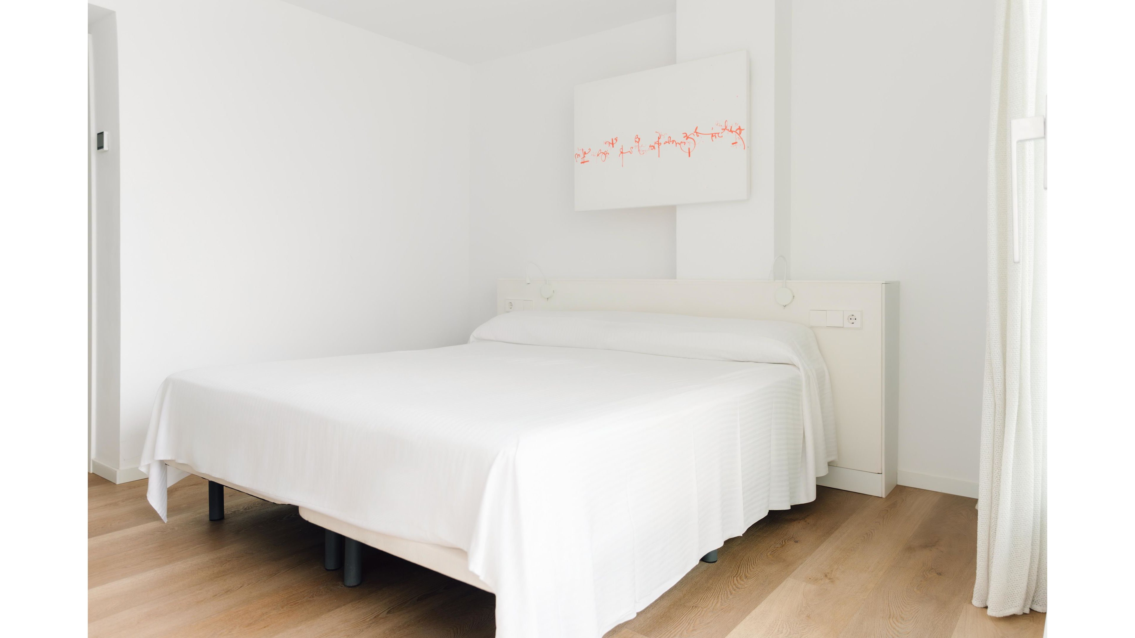 A bright, minimalist bedroom features a neatly made white double bed against a plain wall. Light wood flooring contrasts softly with the white walls, bedding, and headboard. A simple abstract artwork with red accents hangs above the bed, adding a subtle touch of color. Natural light enters from a window on the right, creating a clean and serene atmosphere.