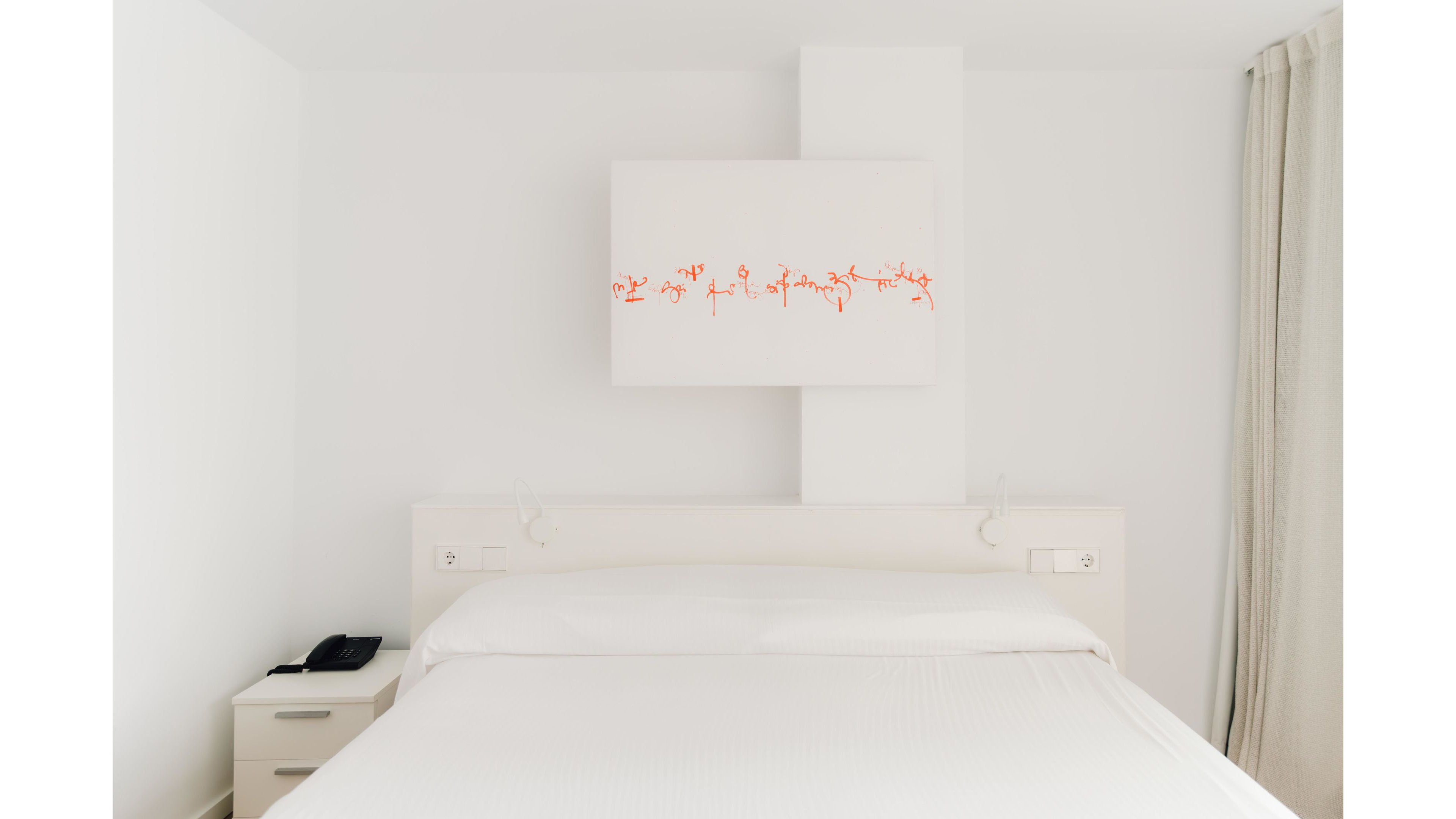 A bright, minimalist bedroom features a large white bed centered against a white wall. Above the headboard hangs a rectangular abstract painting with red accents, adding a subtle pop of color. A small bedside table with a black telephone sits to the left, while light curtains frame the right side of the scene. The overall space appears clean, modern, and serene with a predominantly white palette.