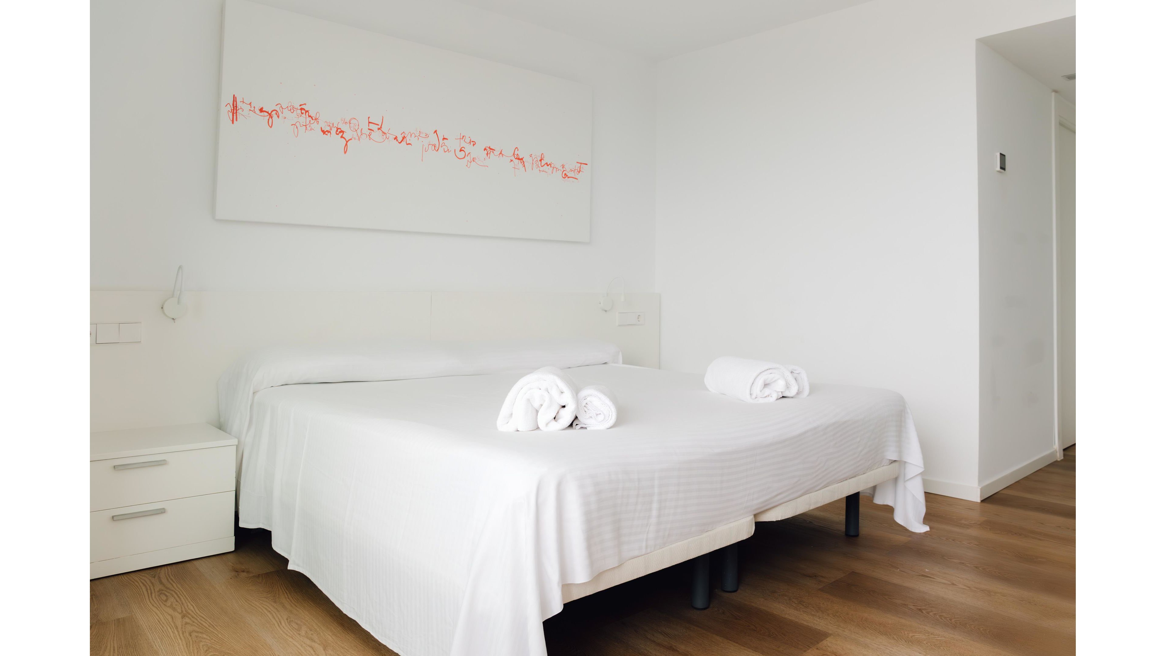 Bright minimalist bedroom featuring a large double bed with crisp white linens. Two neatly rolled white towels are placed on the bed, suggesting a hotel or guest accommodation setting. The room has light wooden flooring, white walls, and a simple bedside table. A modern abstract artwork with red accents hangs above the bed, adding a subtle touch of color to the clean, serene space.
