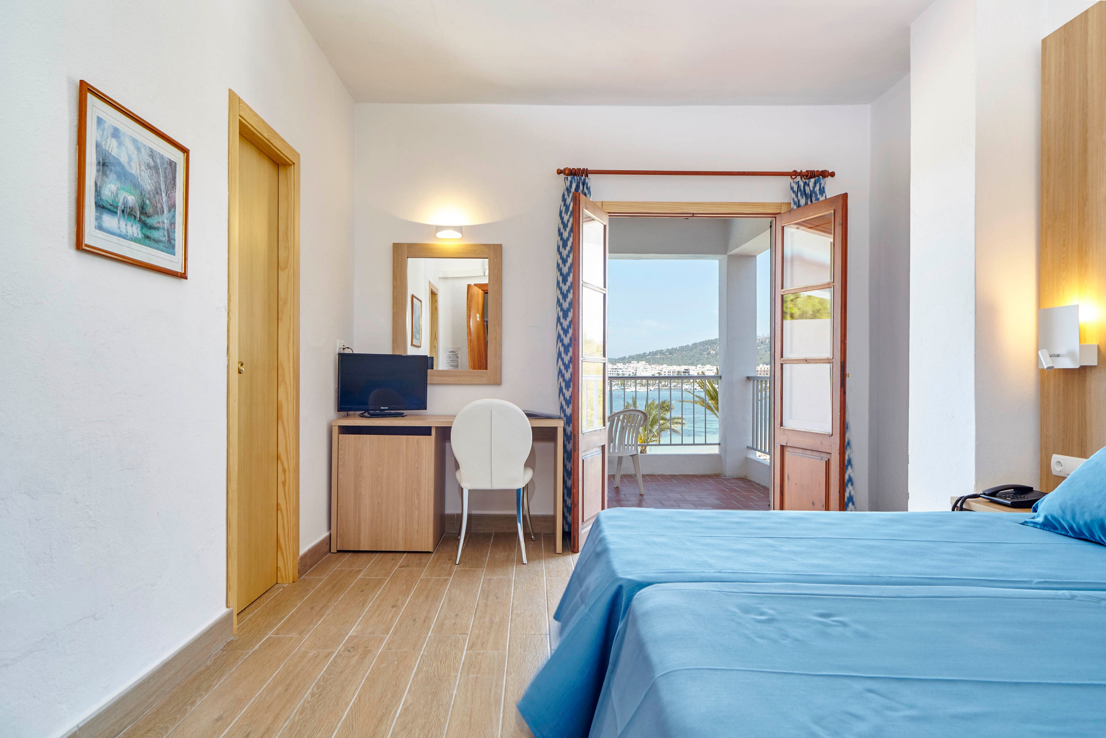 A tidy hotel room features a neatly made bed with blue linens and a small work desk with a computer monitor. Wooden floors and light-colored walls create an airy, minimalist atmosphere. Glass doors open onto a balcony with a view of the sea and distant shoreline. Natural light fills the space, emphasizing the relaxing coastal setting.