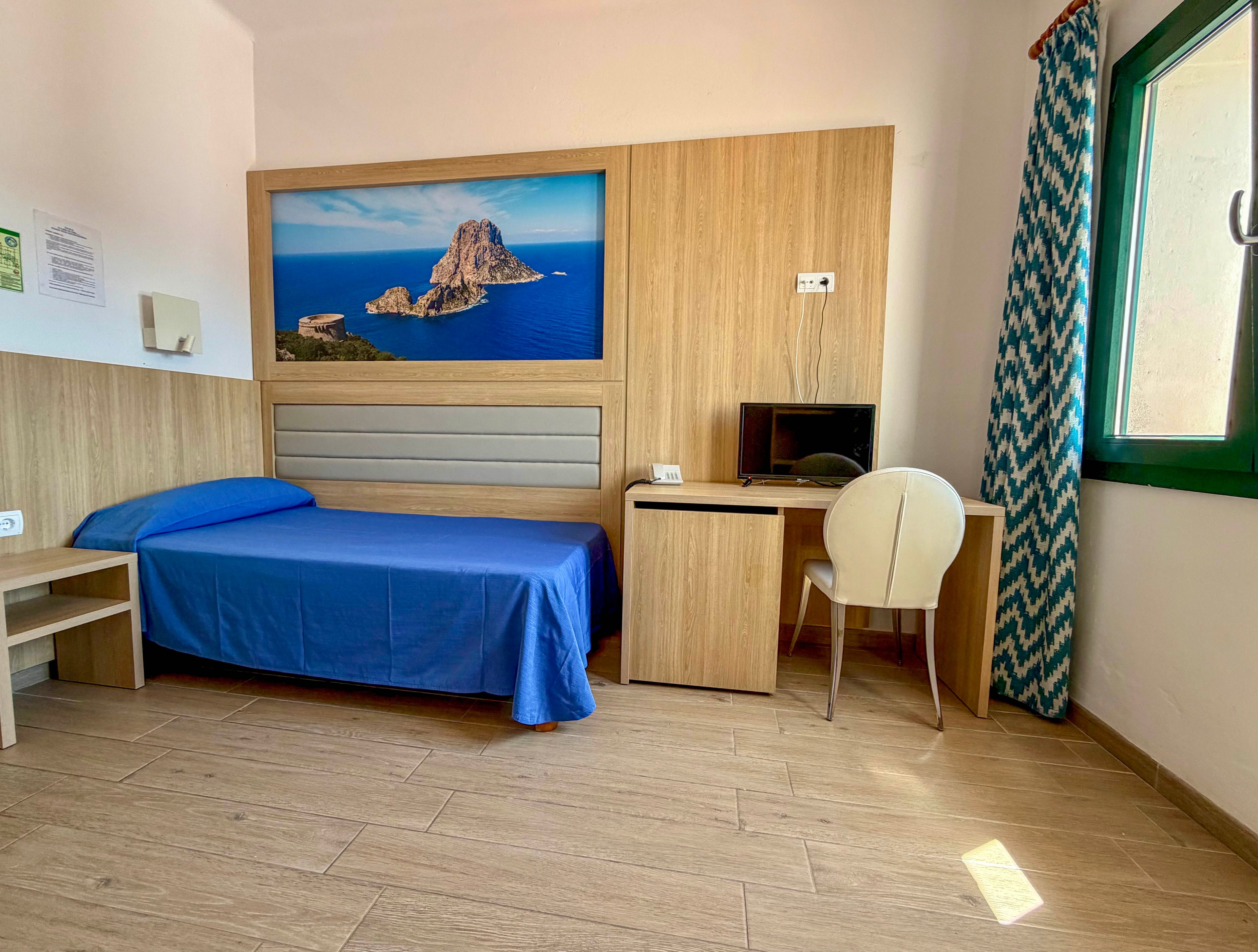 A small hotel room features a single bed with a bright blue bedspread against a wooden paneled wall. A large framed photograph of a rocky island over blue sea hangs above the bed, adding a coastal touch. On the right side, a compact wooden desk with a computer monitor and a white chair sits near a window with patterned blue curtains. Sunlight streams through the window, illuminating the light wood floor and creating a clean, airy atmosphere.