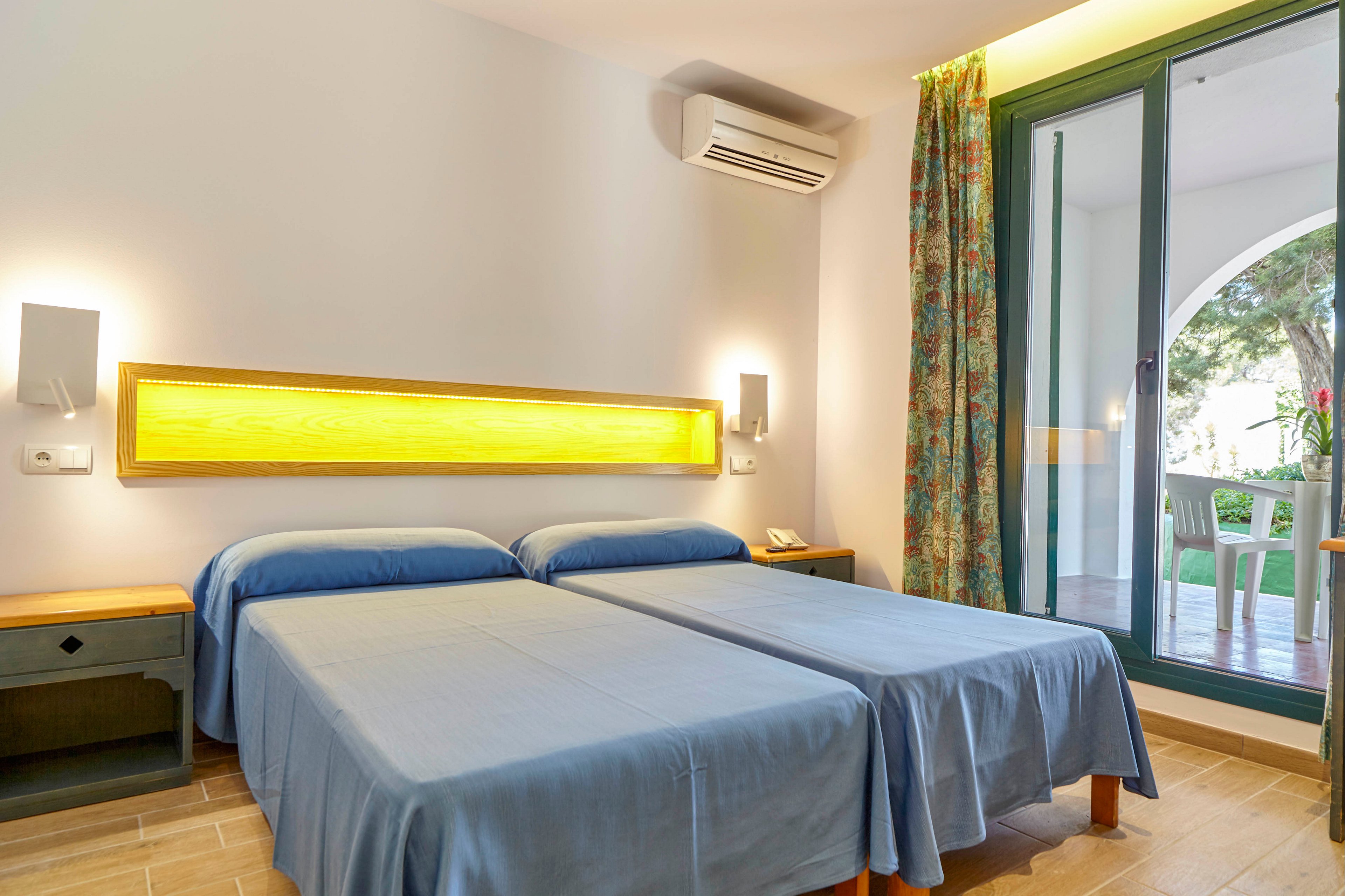A clean hotel room features two neatly made twin beds with blue bedspreads. A warm yellow backlit headboard illuminates the white wall and creates a cozy atmosphere. The room includes bedside tables, wall-mounted reading lights, and an air conditioning unit above the beds. Large glass doors open to a balcony with outdoor chairs and a view of greenery.