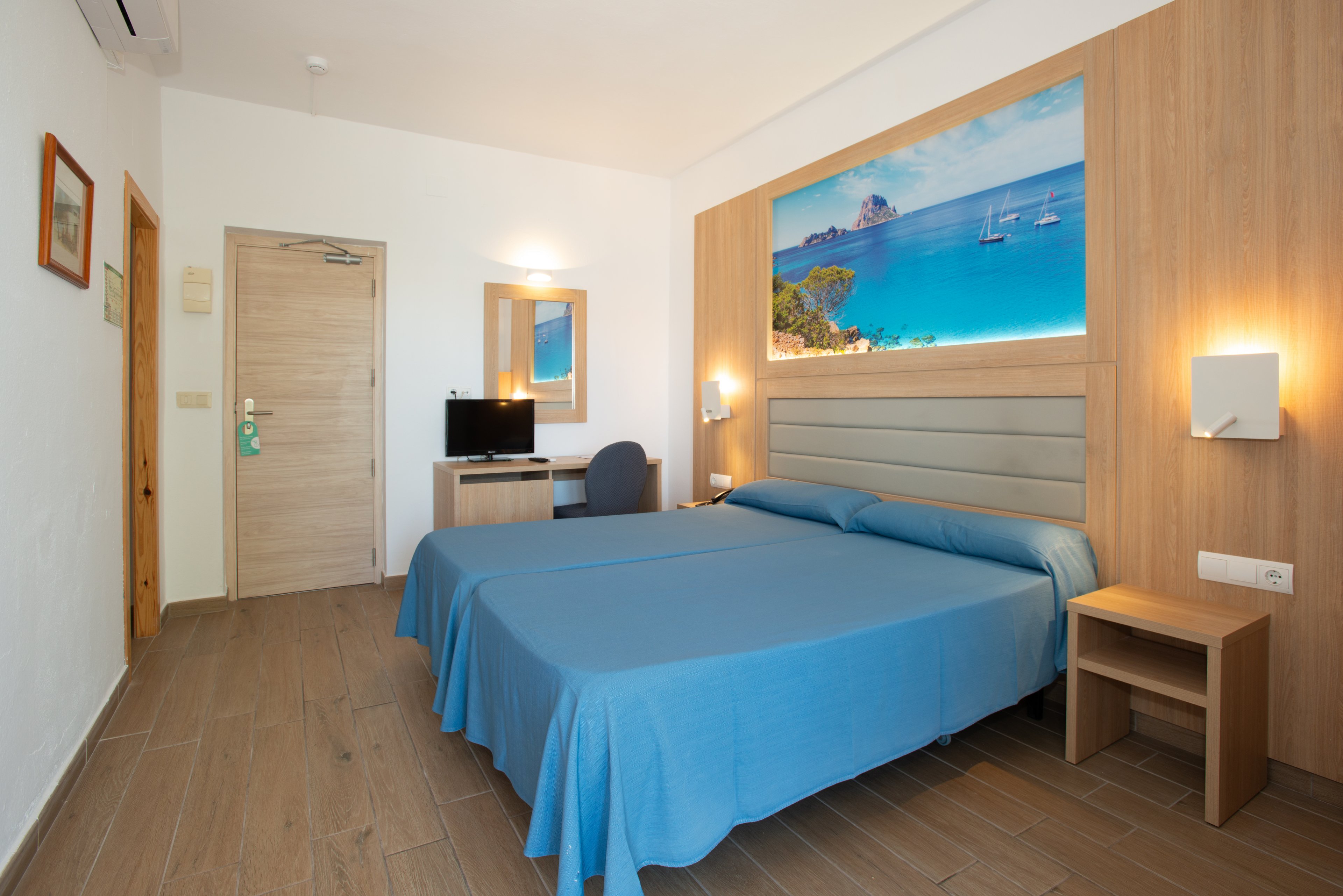 Neat hotel room featuring a double bed with blue bedding is shown. The space includes wooden furniture, a small desk with a computer monitor, and a wall-mounted mirror. A large beach and ocean artwork above the bed adds a coastal theme to the room. Warm lighting and light-colored walls create a clean, inviting atmosphere.