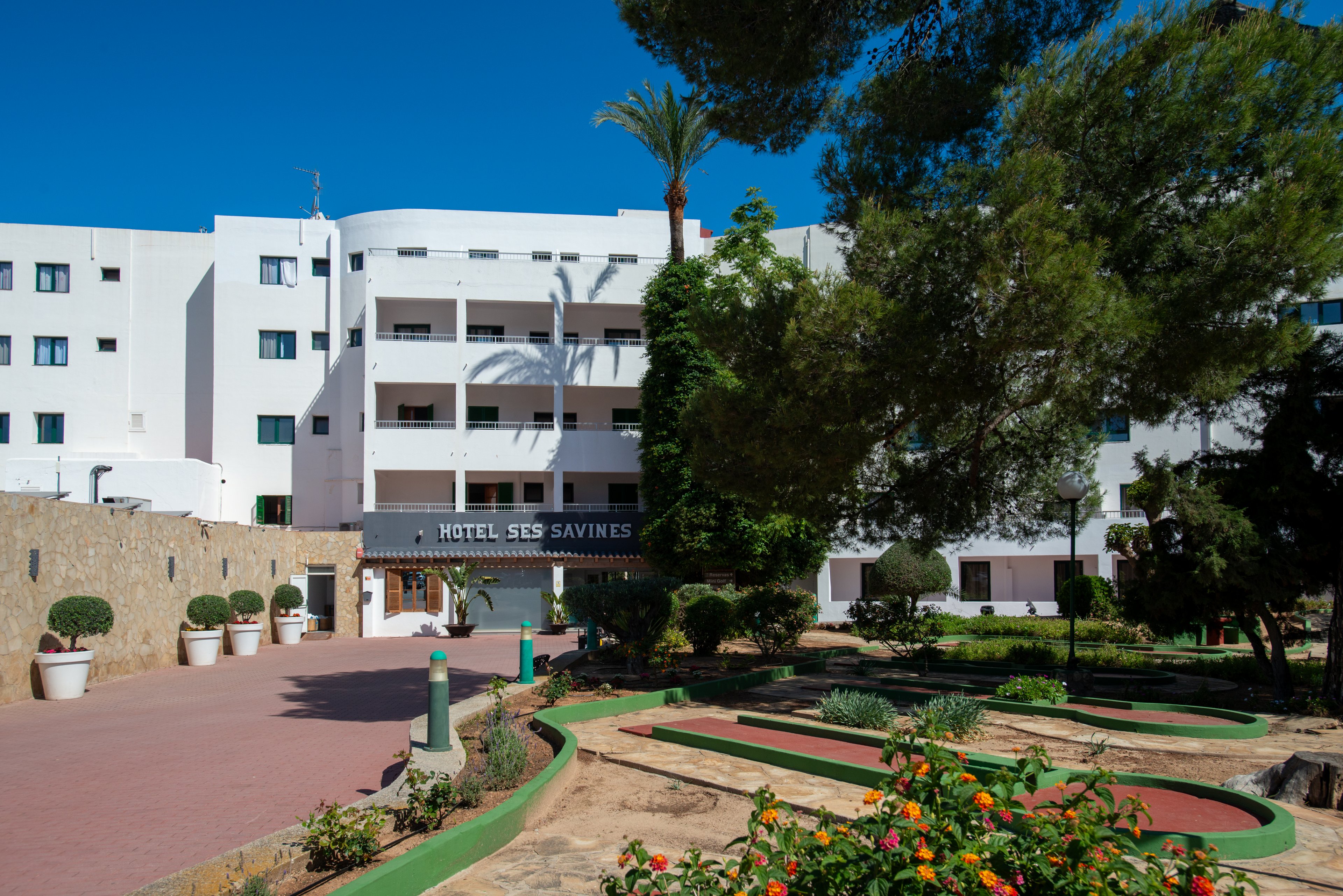 A multi-story white hotel building labeled "Hotel Ses Savines" stands behind a landscaped garden area. The foreground features mini-golf style putting greens bordered in green, sandy paths, and flowering plants. Tall trees frame the scene, casting shadows on a sunny day with clear blue sky. The overall atmosphere is relaxed and inviting, suggesting a holiday or resort setting.
