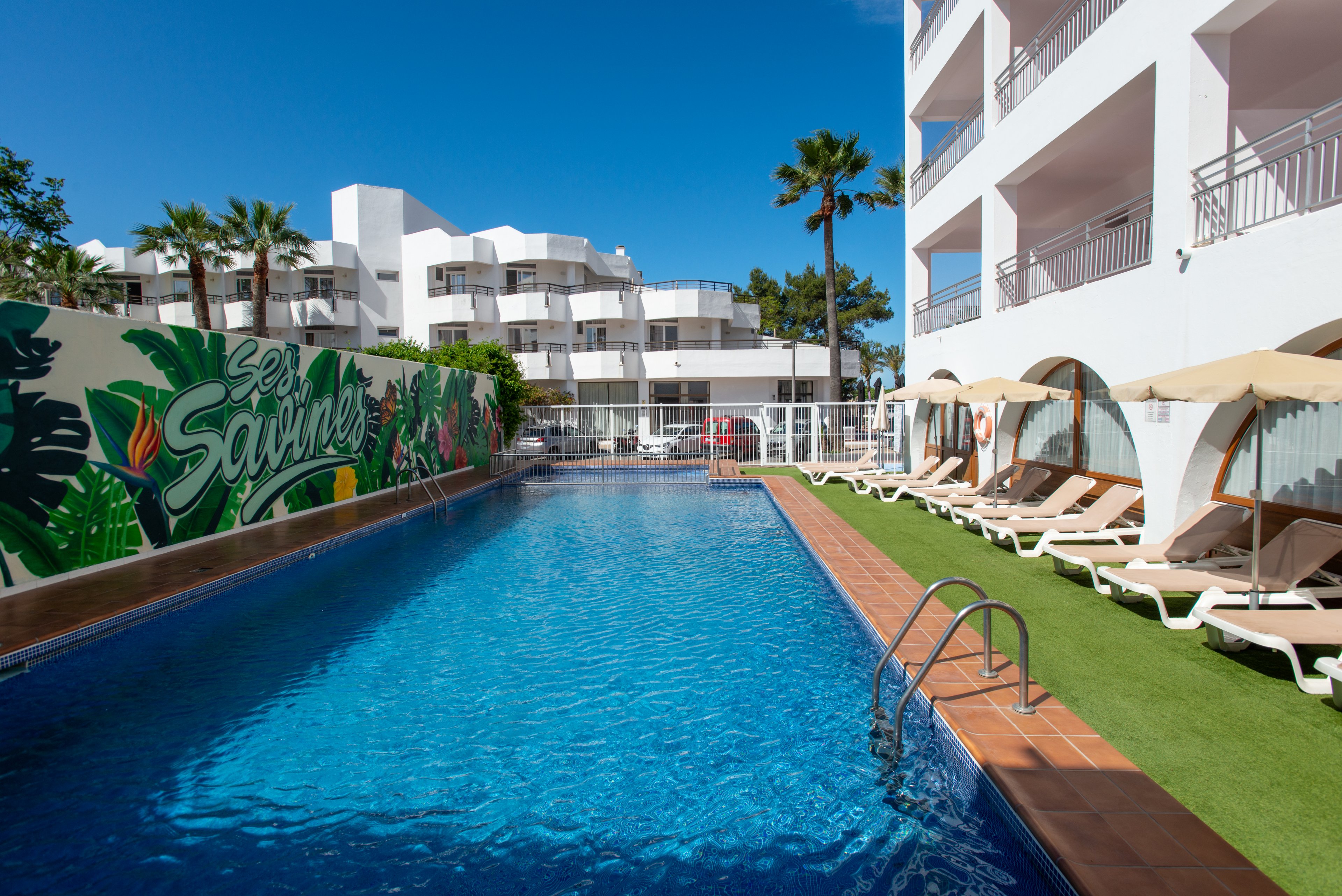Outdoor hotel swimming pool surrounded by modern white buildings under a clear blue sky. Sun loungers with umbrellas line the artificial grass deck along one side of the pool. A colorful mural with tropical leaves decorates the wall at the far end. Tall palm trees add to the relaxed, vacation atmosphere.