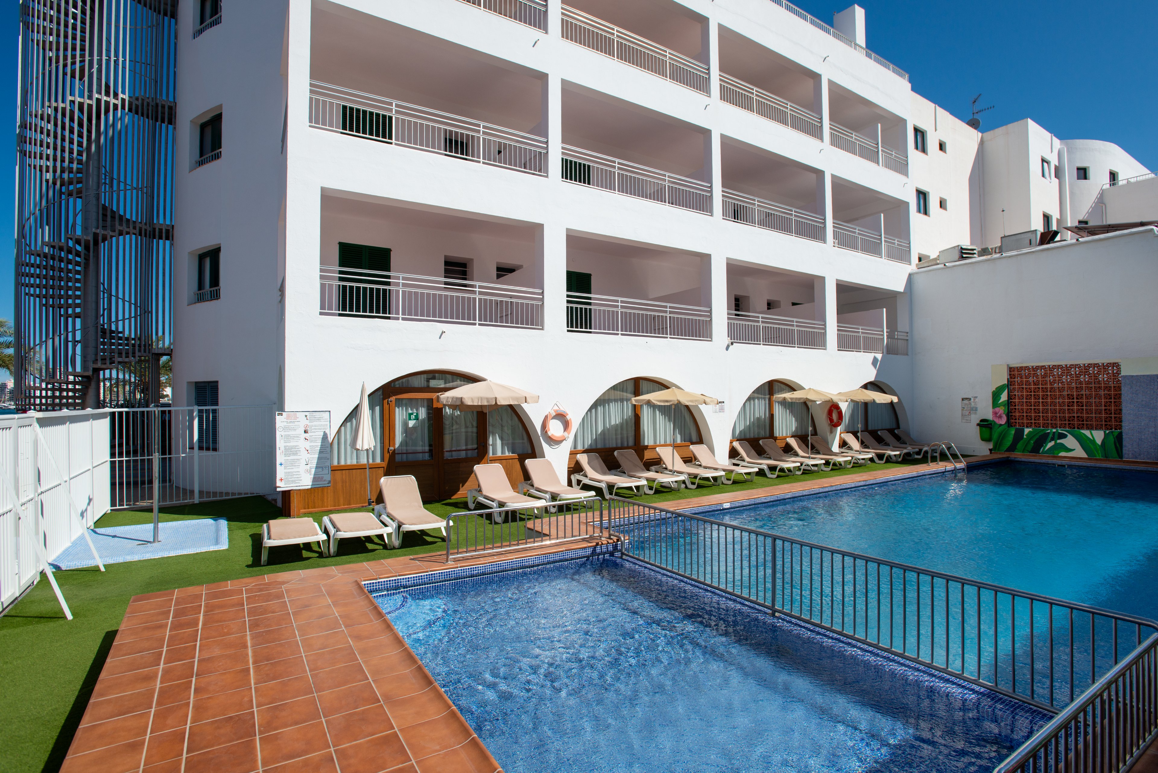 A modern white hotel building overlooks a clear blue outdoor swimming pool. Several sun loungers with umbrellas are arranged along the poolside on artificial grass. The multi-story structure features balconies and arched ground-floor openings facing the water. Bright daylight and clear skies create a relaxed vacation atmosphere. A small fenced area and adjacent structures suggest additional recreational facilities nearby.