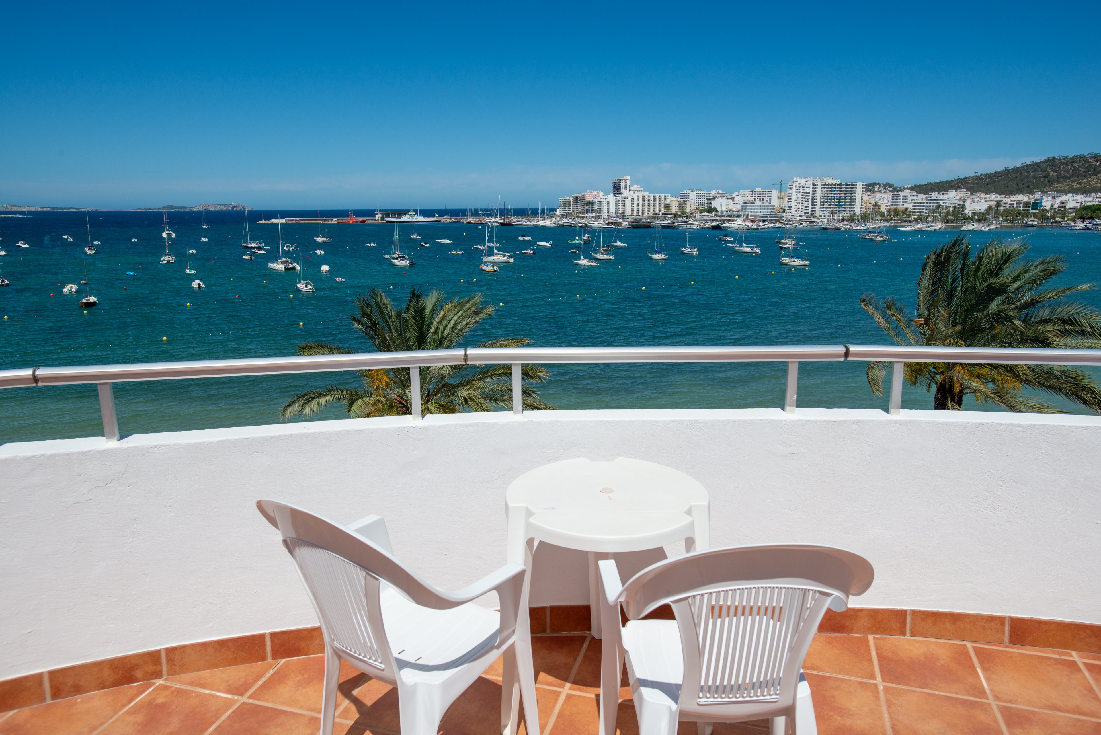 A curved balcony with terracotta tiles features a small round white table and two white plastic chairs. The balcony overlooks a calm blue sea filled with anchored boats and yachts. In the distance, a coastal city with white buildings lines the shoreline beneath a clear blue sky. Palm trees rise near the water, adding a tropical feel to the scenic waterfront view.