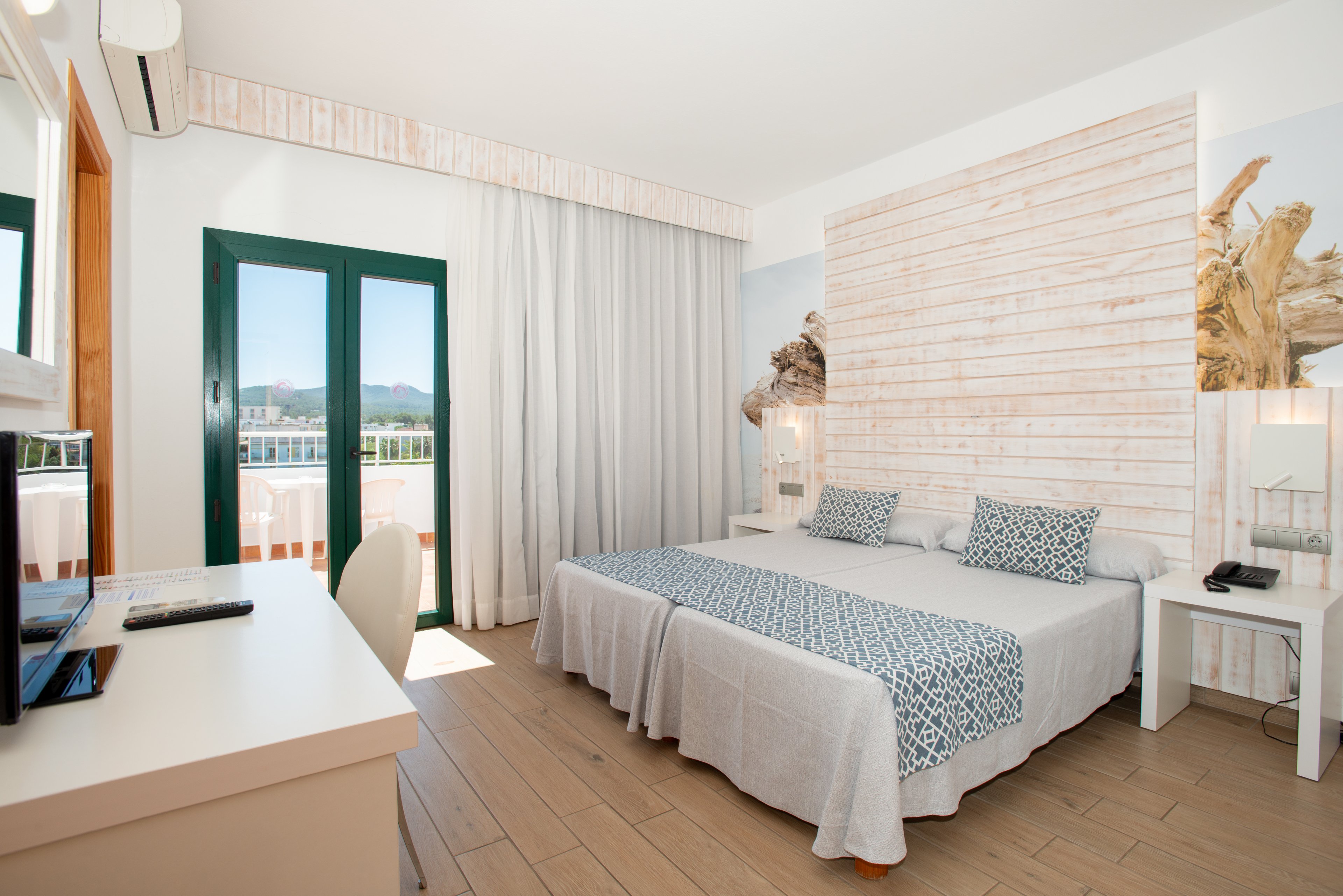 A bright, modern hotel bedroom features two single beds pushed together with white linens and patterned blue runners. The room is decorated in a light coastal style with a white wood accent wall and neutral flooring. Large green-framed glass doors open to a balcony with a scenic mountain and water view. A desk with a computer monitor and chair sits opposite the beds, and bedside tables with lamps complete the cozy, minimalist space.