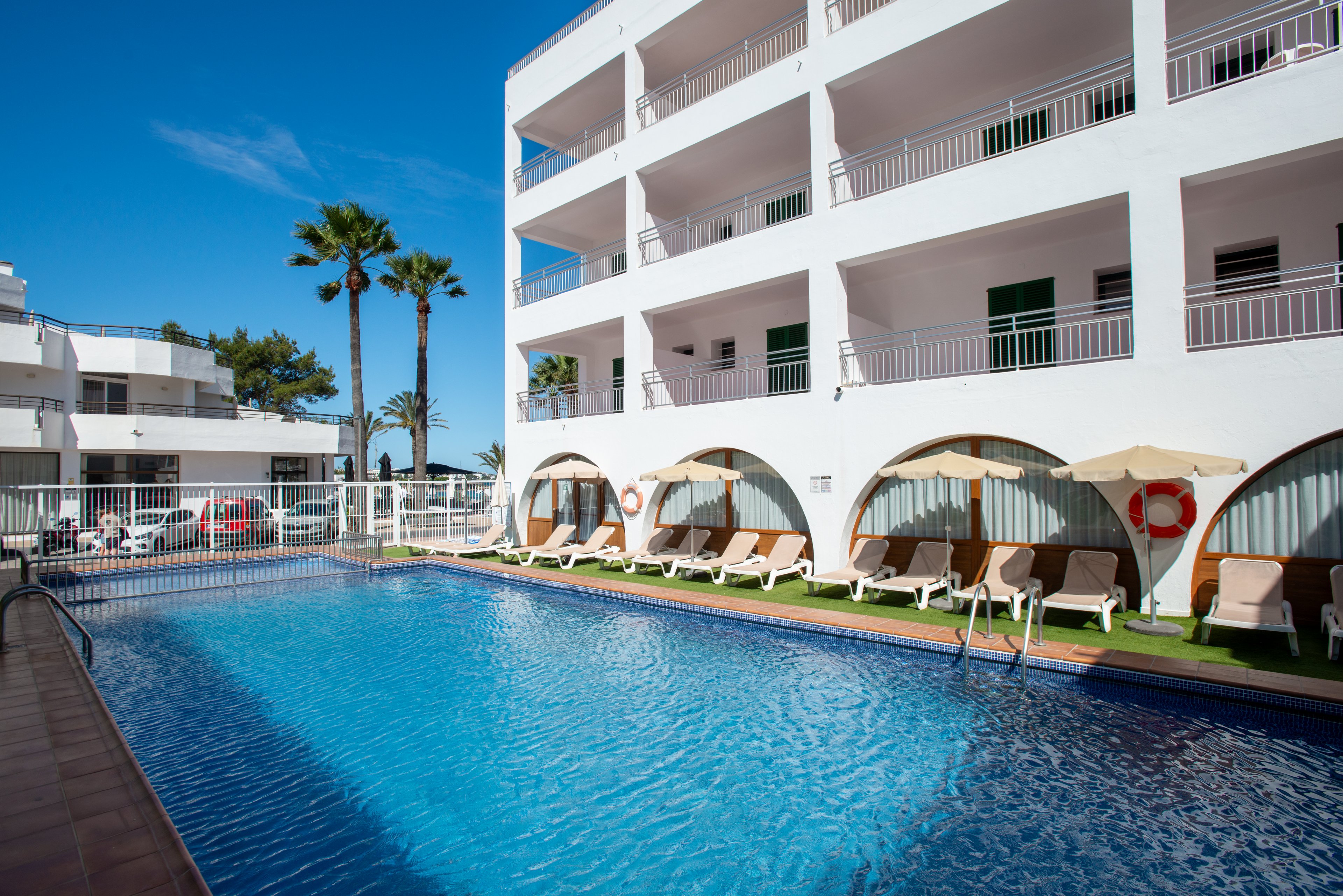An outdoor swimming pool sits beside a white multi-story hotel building under a clear blue sky. Sun loungers and umbrellas line the poolside area, some placed under arched alcoves on artificial grass. Palm trees and additional white buildings are visible in the background, enhancing the resort atmosphere. The scene suggests a warm, sunny day ideal for relaxing by the water.