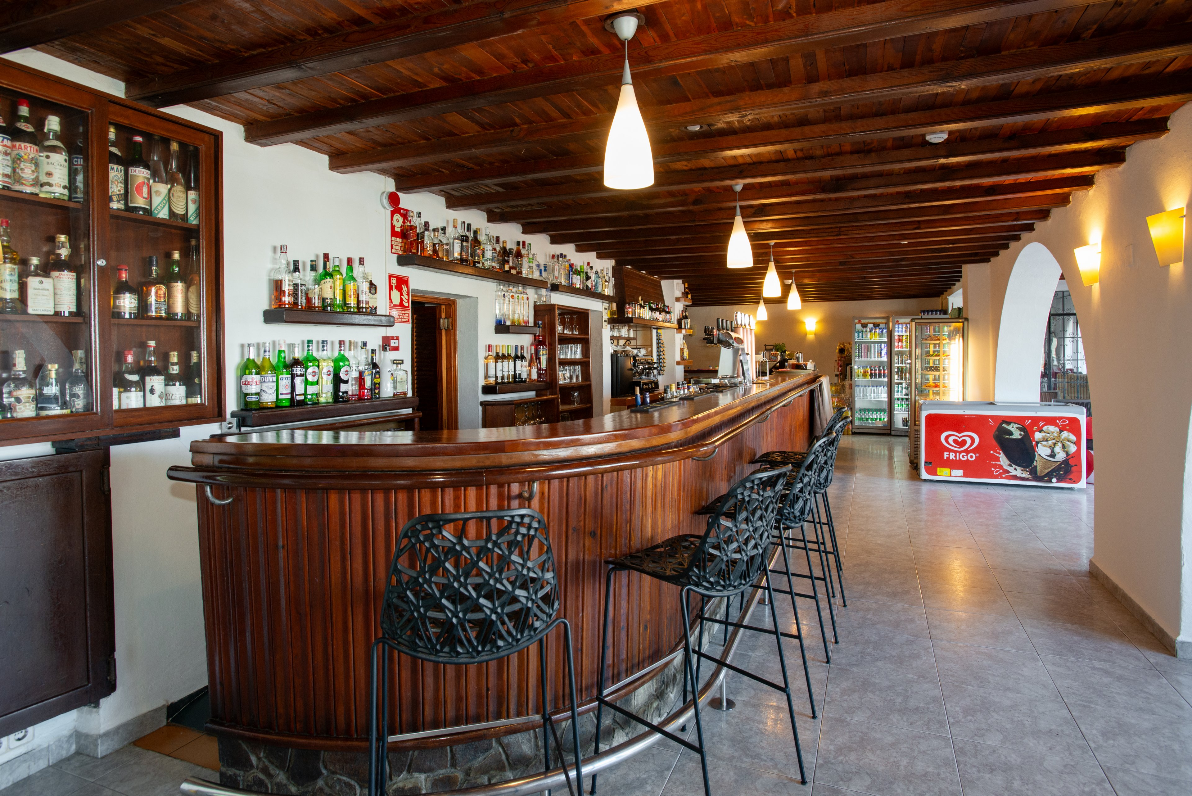 An inviting bar interior features a long curved wooden counter lined with modern black bar stools. Bottles of liquor and spirits are neatly arranged on shelves behind the bar, with warm pendant lights hanging from the wooden ceiling. The tiled floor leads toward a refrigerated display and ice cream freezer at the far end of the room. The space is empty of people, emphasizing the clean, organized, and welcoming atmosphere.