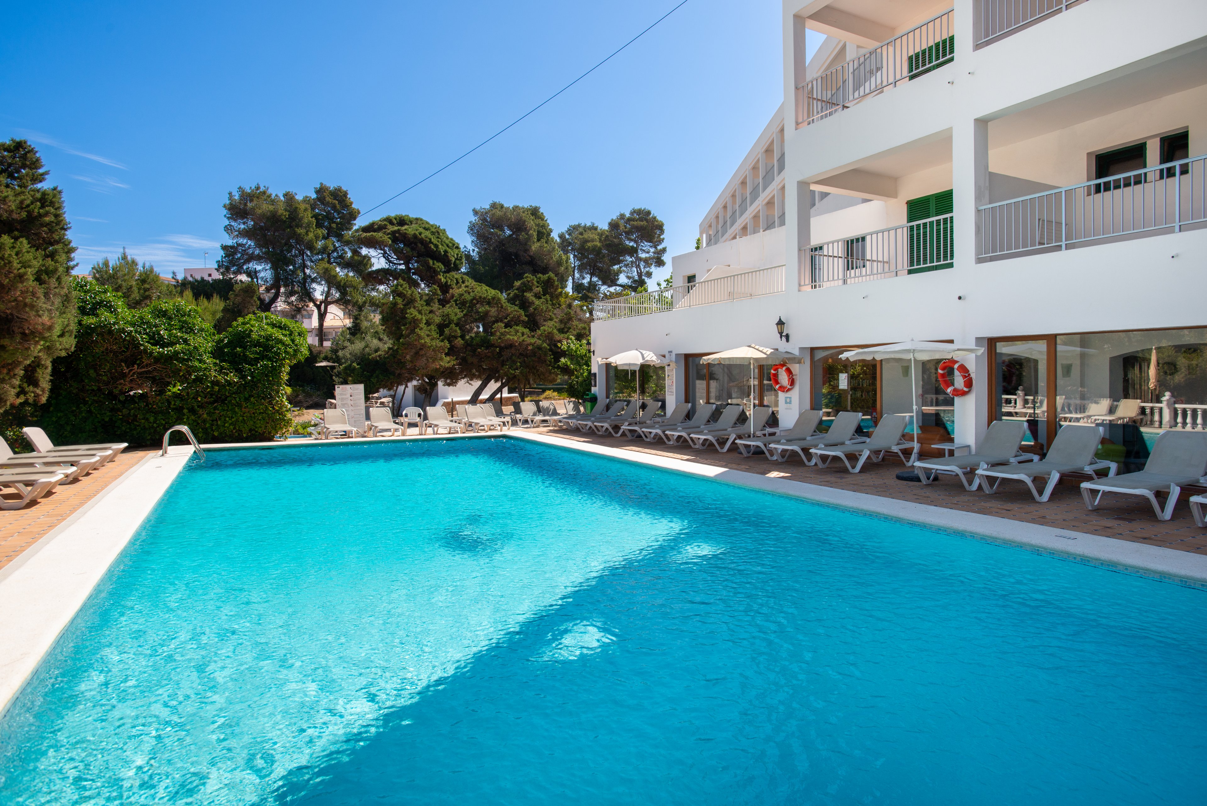 An outdoor swimming pool stretches alongside a modern white hotel building. Numerous sun loungers line the pool deck, some shaded by large umbrellas. Lush green trees and shrubs surround the area, contrasting with the bright blue water and clear sky. The scene suggests a relaxing vacation setting at a resort or coastal hotel.