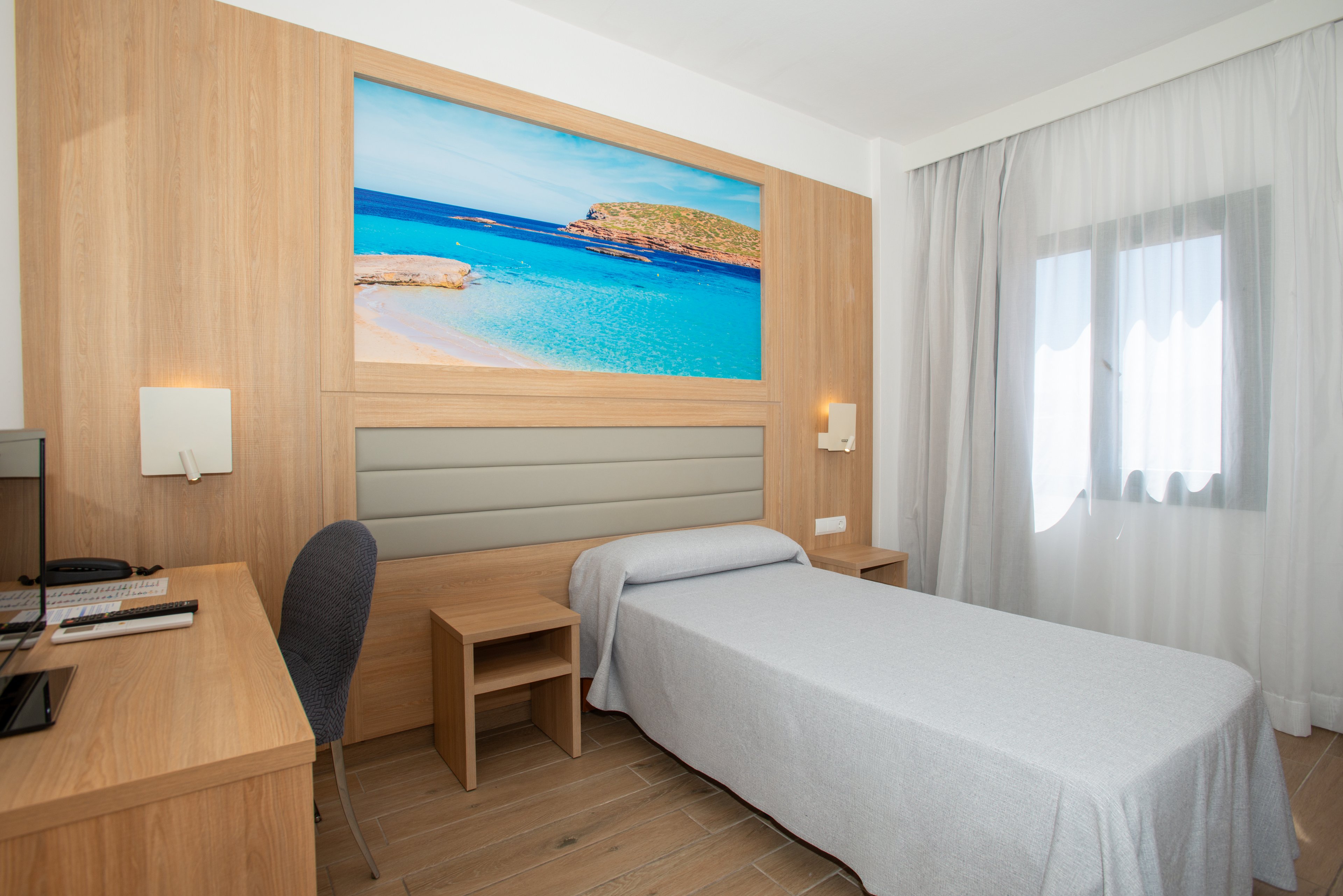 Modern hotel room featuring a neatly made single bed with a light gray bedspread. The space includes a wooden desk with a chair, computer monitor, keyboard, and telephone. A large wall art print of a tropical beach scene adds vibrant color above the headboard. Sheer white curtains cover the window, allowing soft natural light to fill the clean, minimalist interior.