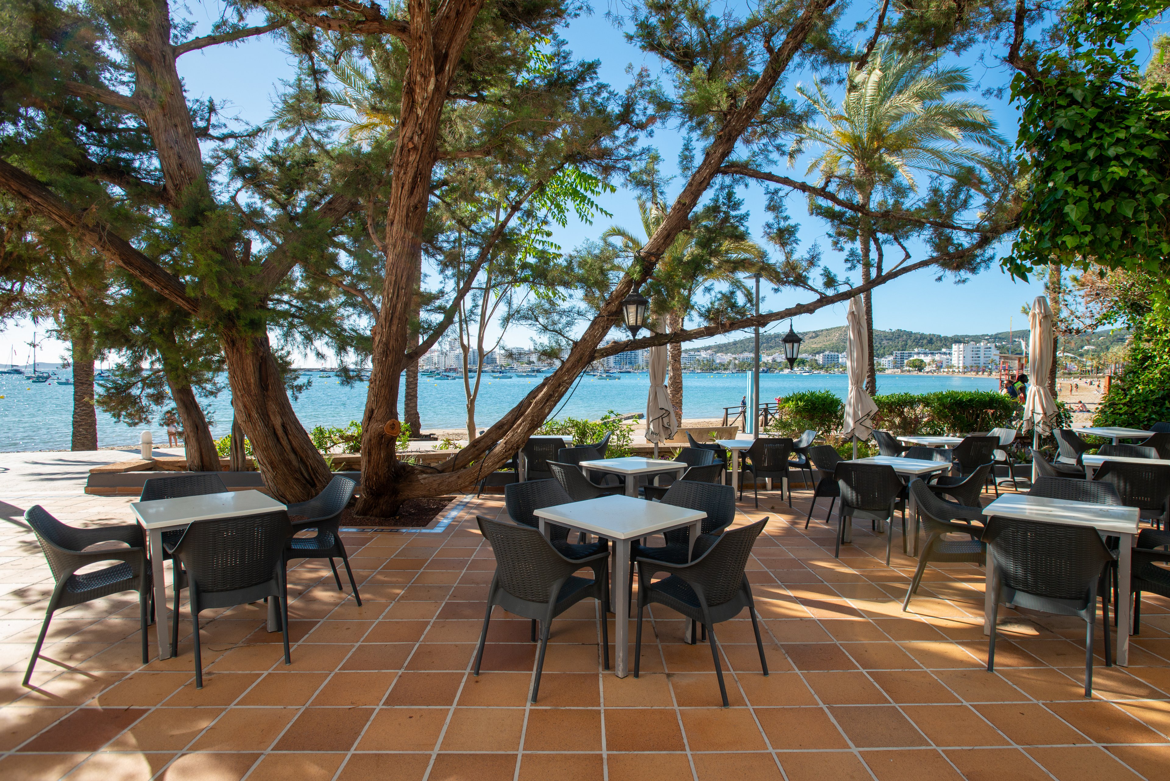 An outdoor cafe terrace features multiple empty tables and chairs arranged on a terracotta-tiled floor. Large trees and palm trees provide shade and frame the view of the water. In the background, a calm bay with boats and distant buildings suggests a coastal town or resort. The bright daylight and clear blue sky create a relaxed, vacation-like atmosphere.