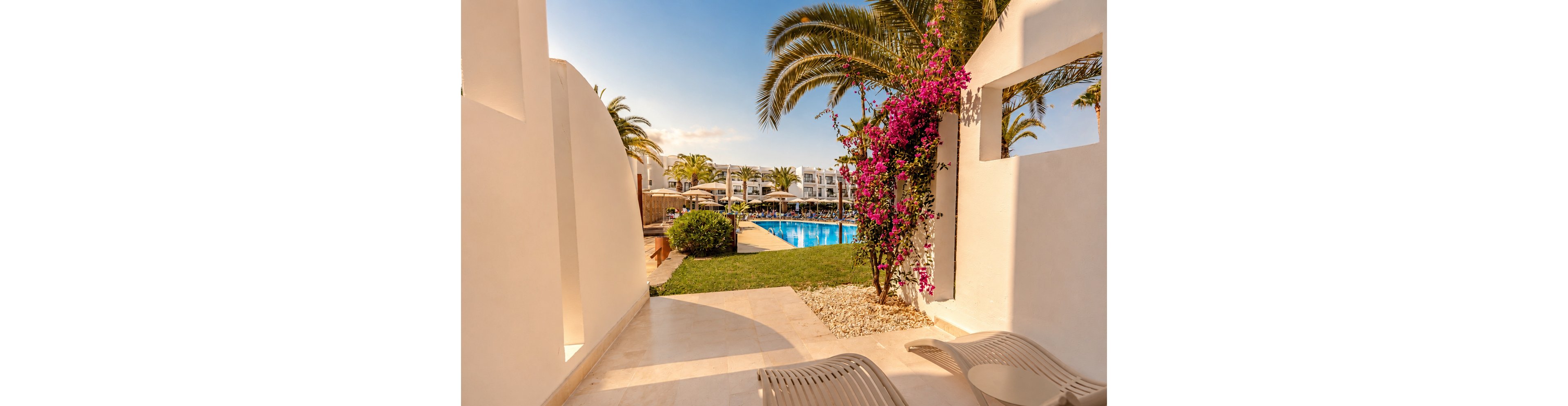 A bright resort terrace opens onto a view of a blue outdoor swimming pool. White modern buildings and sun loungers surround the pool under clear skies. Palm trees and vibrant pink bougainvillea add a tropical touch to the scene. The warm sunlight and clean architectural lines create a relaxing vacation atmosphere.