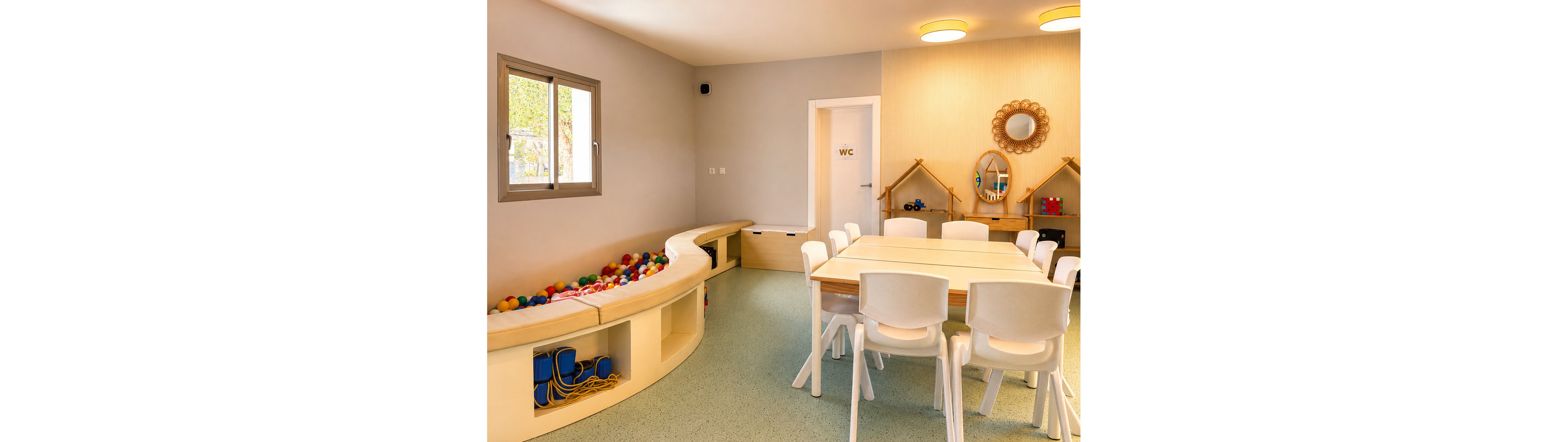 A modern children’s playroom features a central white table surrounded by small chairs. Along one wall is a curved built-in ball pit filled with colorful balls and storage cubbies holding toys. The room includes a door marked with the text "WC" and a cozy play area with small wooden house-shaped shelves and decorative mirrors. Soft lighting and pastel walls create a warm, inviting atmosphere for kids’ activities.