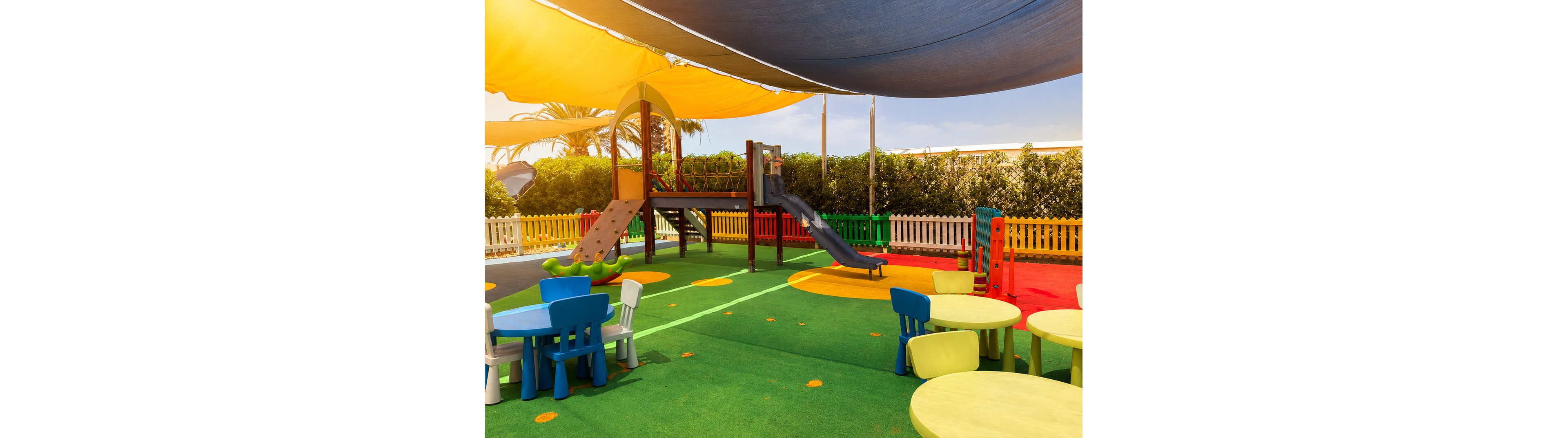 A vibrant outdoor playground features a central play structure with slides and climbing elements on a soft, colorful rubber surface. Round plastic tables and chairs in bright colors are arranged in the foreground, suggesting a child-friendly seating area. Large fabric canopies provide shade over the entire play zone, while a white picket-style fence and greenery enclose the space. The scene is sunlit and cheerful, designed for young children’s recreation and safety.