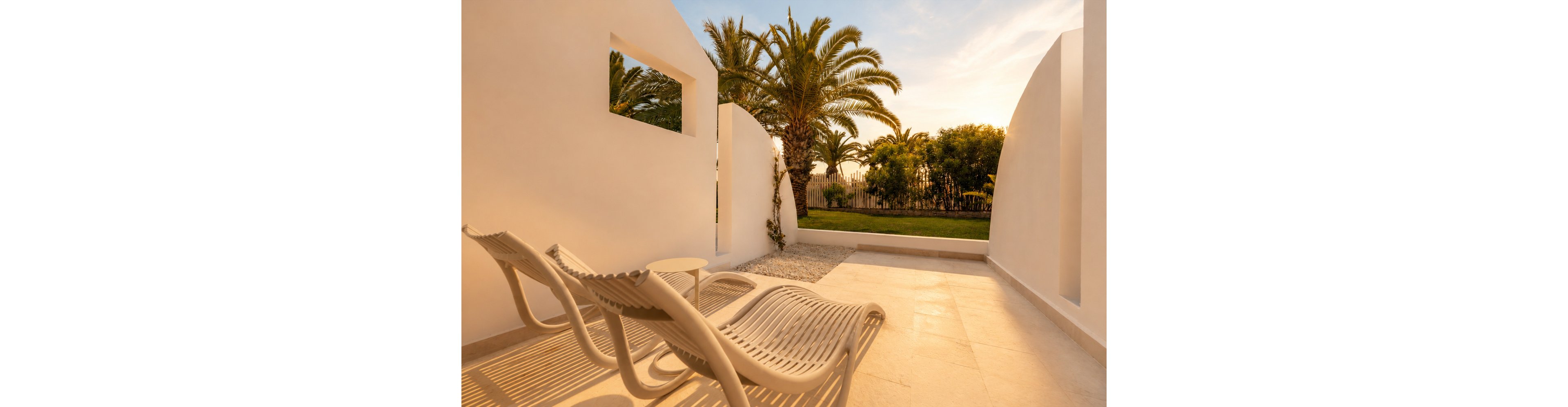 A modern outdoor patio features two sculpted sun loungers facing a small side table. The space is framed by smooth white walls and a minimalist stone floor. In the background, tall palm trees and greenery are softly lit by the warm evening light. Long shadows and a golden sky create a tranquil, resort-like atmosphere.
