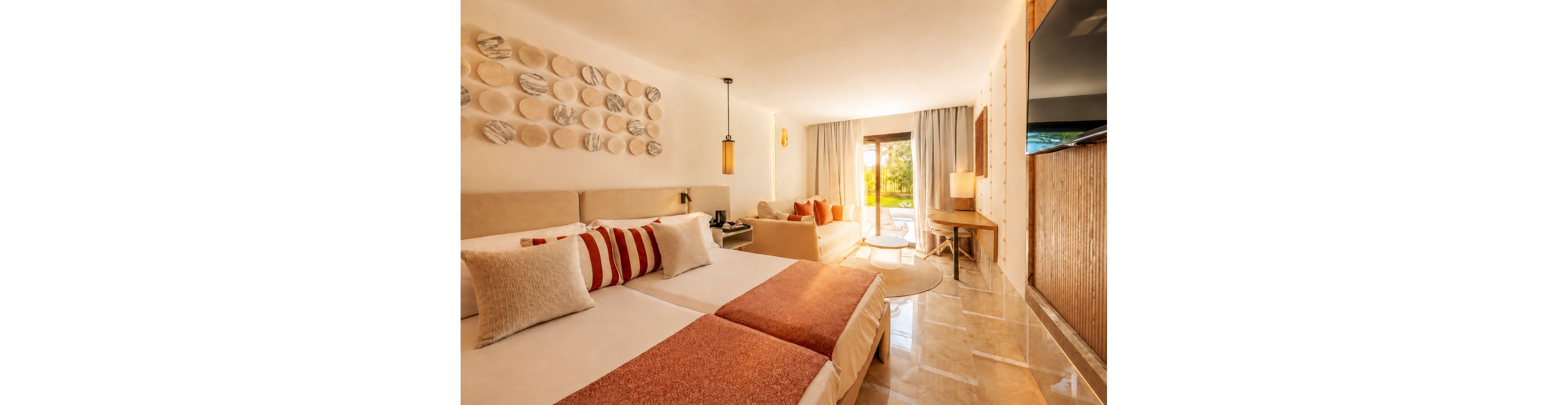 A contemporary hotel room features two neatly made beds with red and white accent pillows and throws. Warm lighting, marble-style wall art, and neutral furnishings create a cozy, inviting atmosphere. A sofa seating area, wooden desk, and floor lamp extend the living space toward large glass doors. The doors open to a sunlit terrace with views of greenery outside, enhancing the airy, resort-like feel.