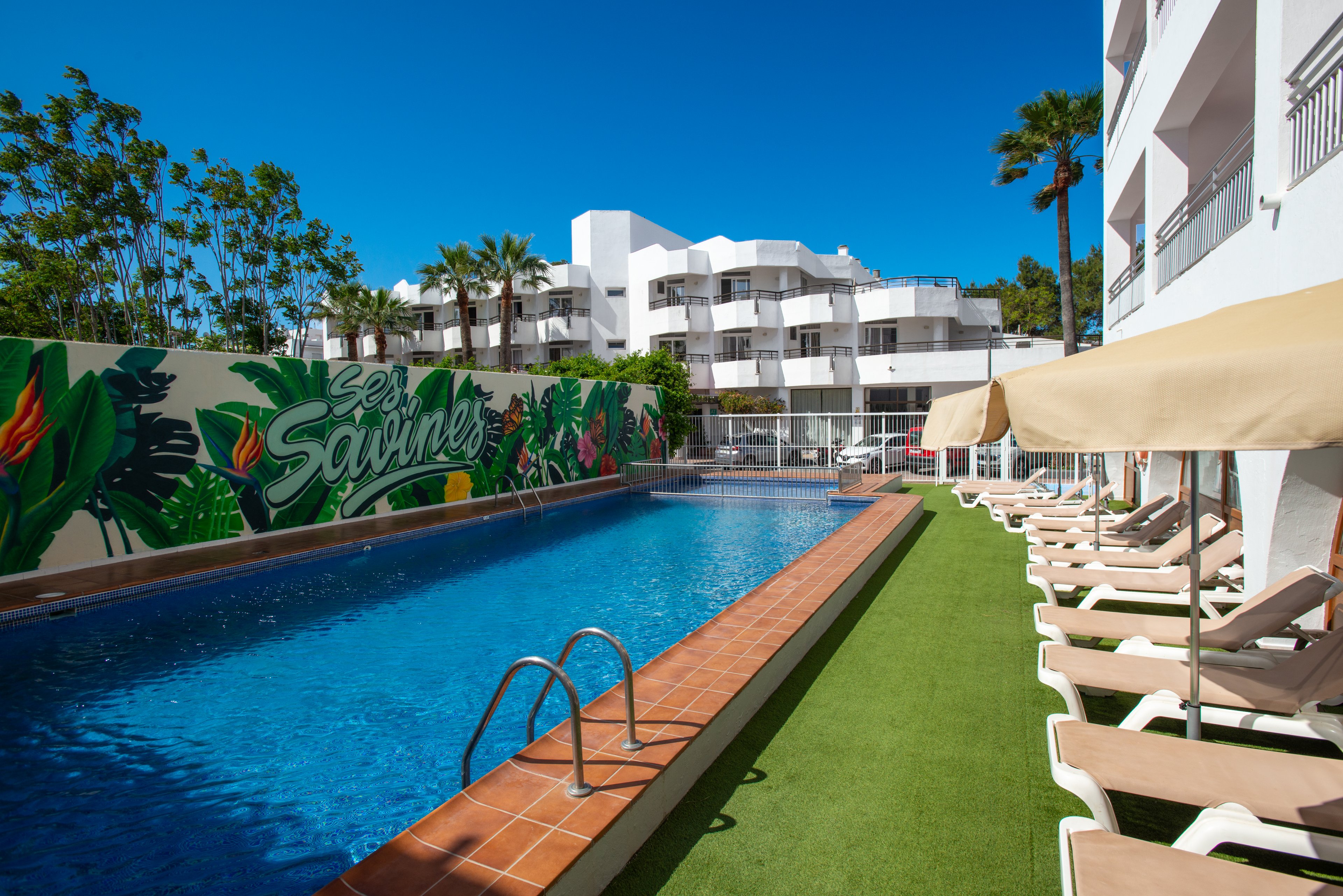 Outdoor hotel swimming pool lined with beige sun loungers sits beside a modern white building under a clear blue sky. A colorful tropical mural with the text "Les Sabines" decorates the wall along one side of the pool. Green artificial grass borders the pool deck, and palm trees rise behind the buildings. The scene suggests a bright, relaxing vacation atmosphere in a warm climate.