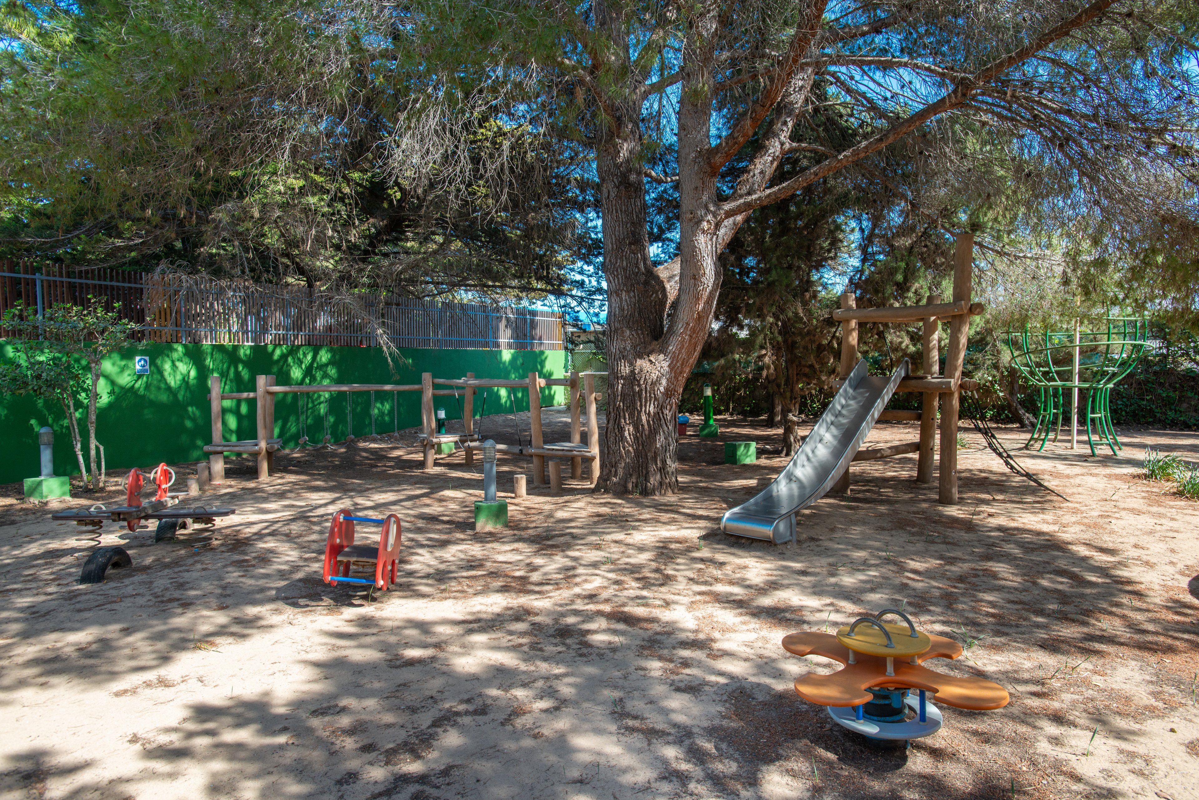 An empty outdoor playground is set beneath tall, shady trees on a sunny day. The play area features a metal slide, wooden climbing structures, and several spring riders on sandy ground. A green fence and wall enclose the space, adding bright color to the natural setting. The scene feels calm and inviting, ready for children to play.