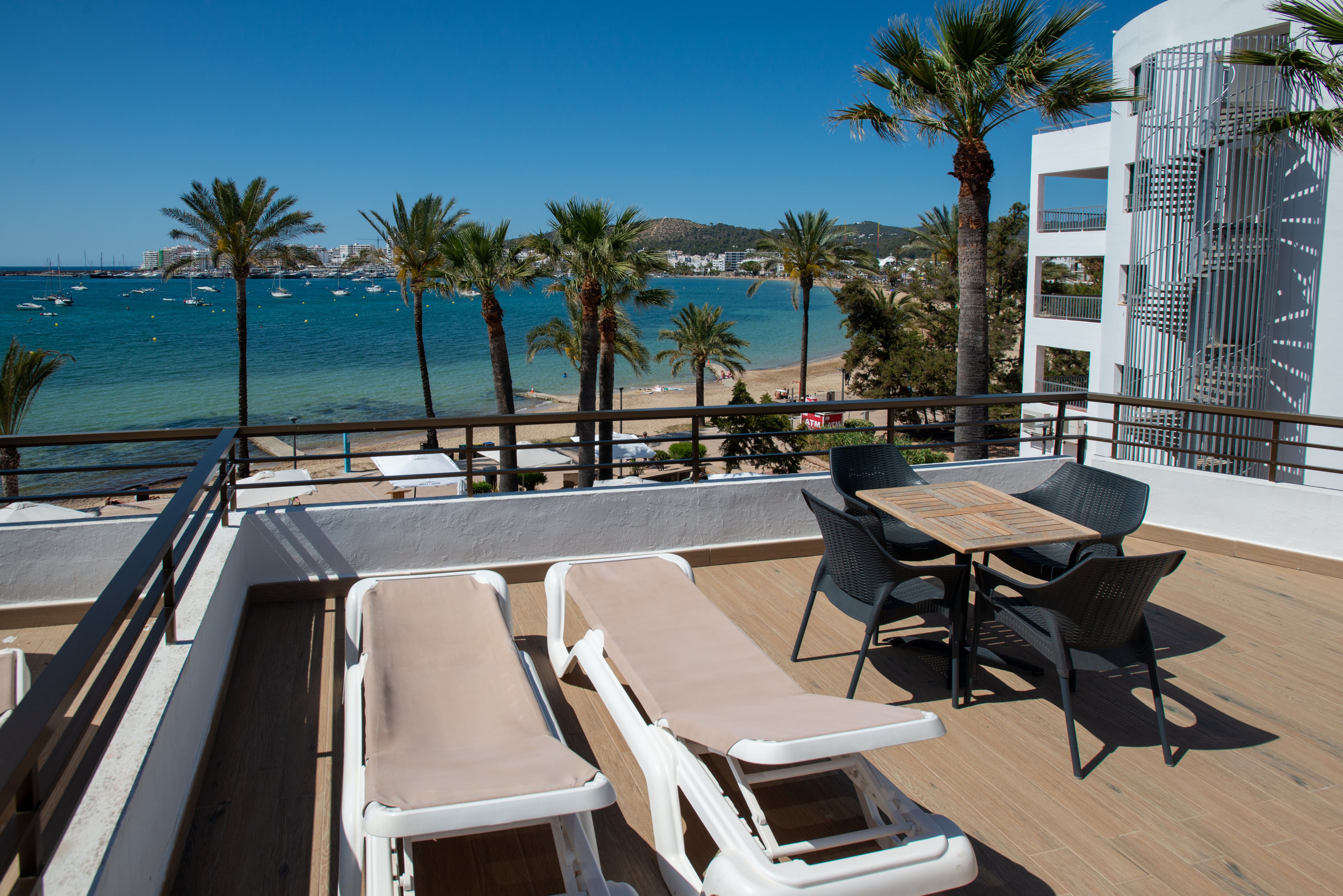 A modern hotel terrace overlooks a bright blue seaside bay. Two sun loungers and a small dining table with four chairs sit on a wooden deck. Palm trees line the beachfront below, and boats are anchored in the calm water. Clear skies and strong sunlight create a relaxed, vacation atmosphere.