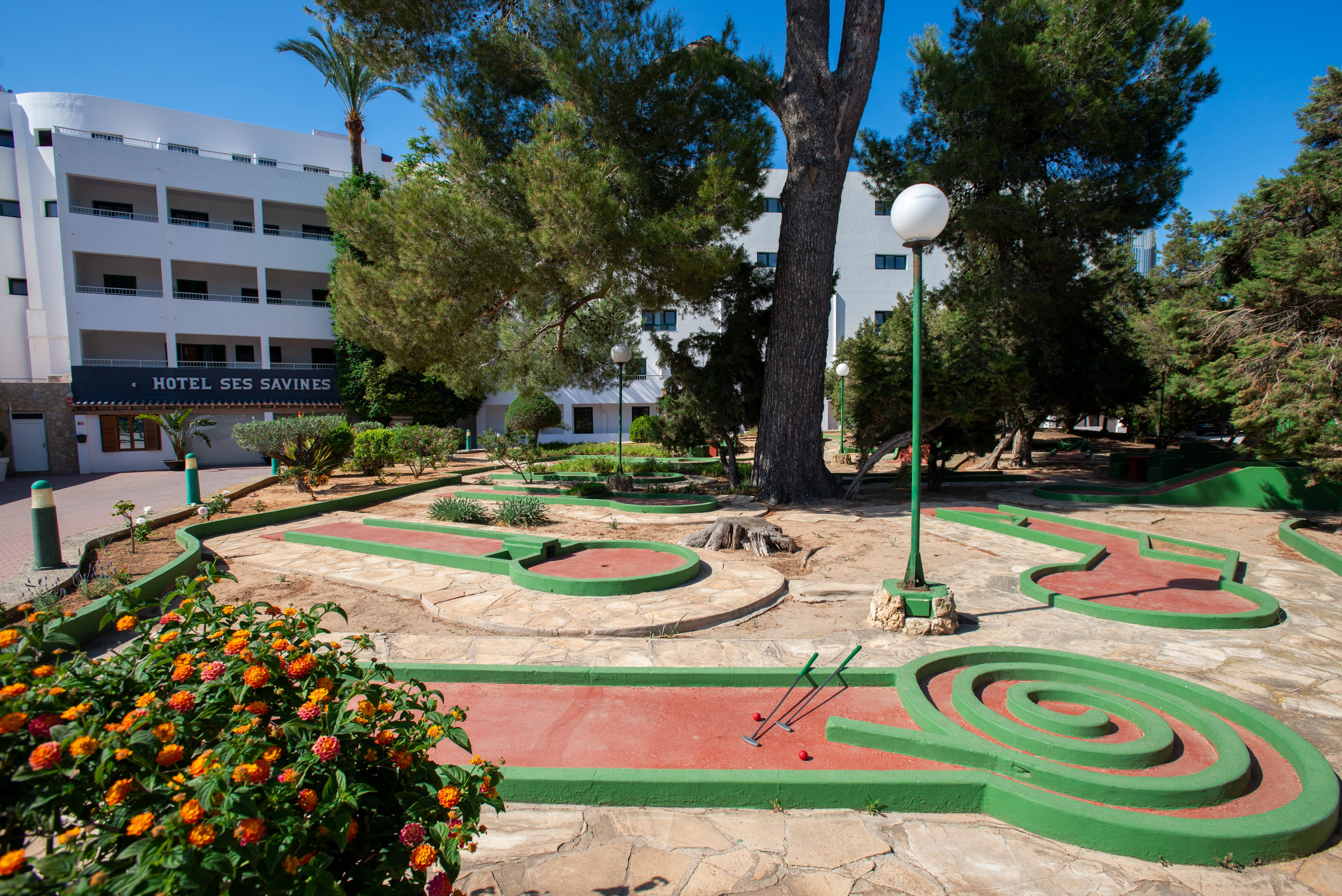An outdoor mini golf course is set in front of a white multi-story hotel building surrounded by tall trees. Curving red and green putting lanes wind across a stone-paved ground, with a spiral obstacle in the foreground. A pair of golf putters and balls rest on one of the lanes, suggesting recent play. Colorful flowers and green posts with spherical lamps add to the relaxed holiday atmosphere. The hotel sign reads "HOTEL SES SAVINES" on the facade.