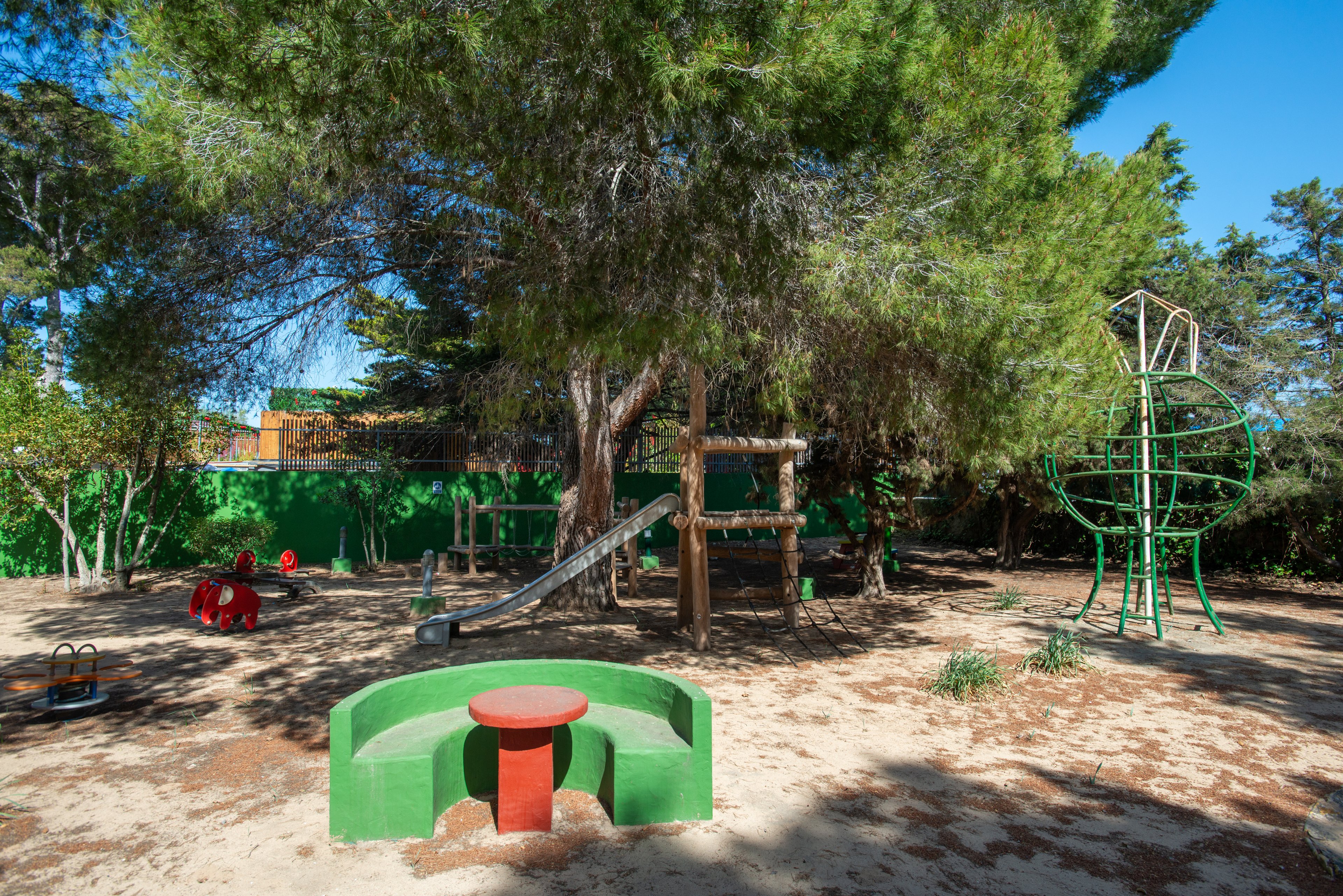 An outdoor playground sits beneath tall, leafy trees on a sunny day. The area features a slide, climbing structures, and spring riders scattered across a sandy ground. A green bench with a small red table stands in the foreground, adding bright color to the scene. The surrounding trees and green fence create a shaded, enclosed play space with no people present.
