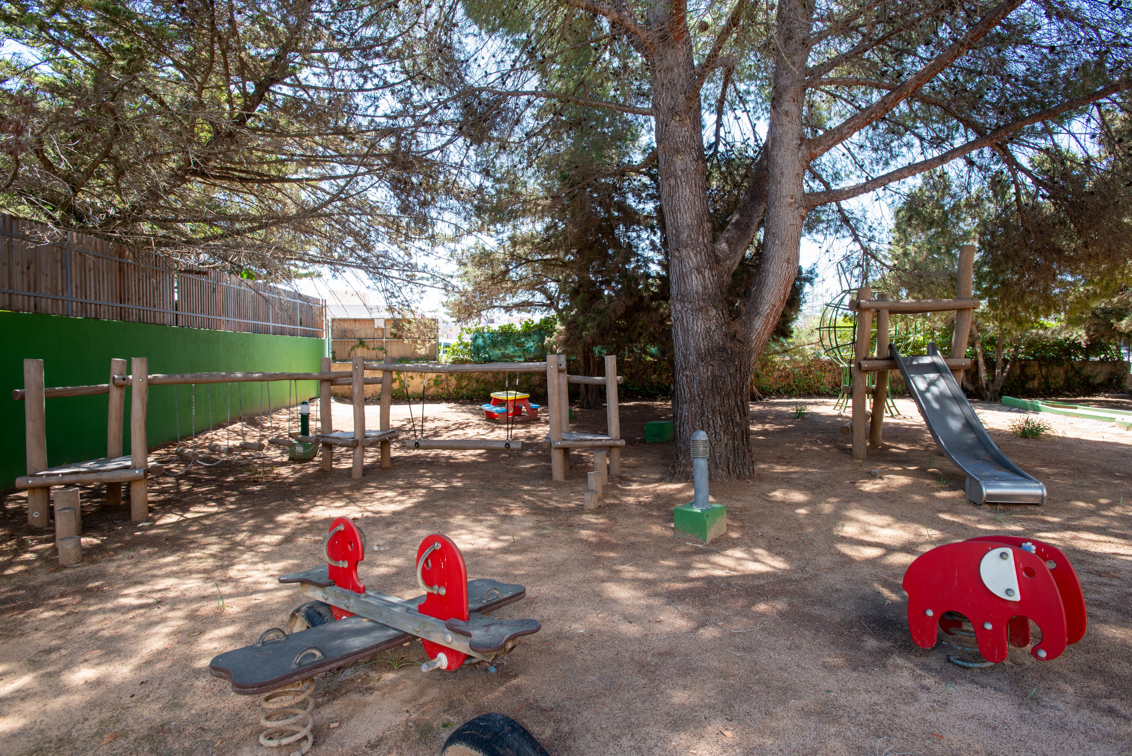 An outdoor playground is set beneath tall trees, providing shade over the play area. The space features wooden climbing structures, a slide, and several spring-mounted rocking toys. Bright red play elements stand out against the sandy ground and natural wood. A green fence and surrounding vegetation enclose the quiet recreational area, suggesting a safe space for children to play.