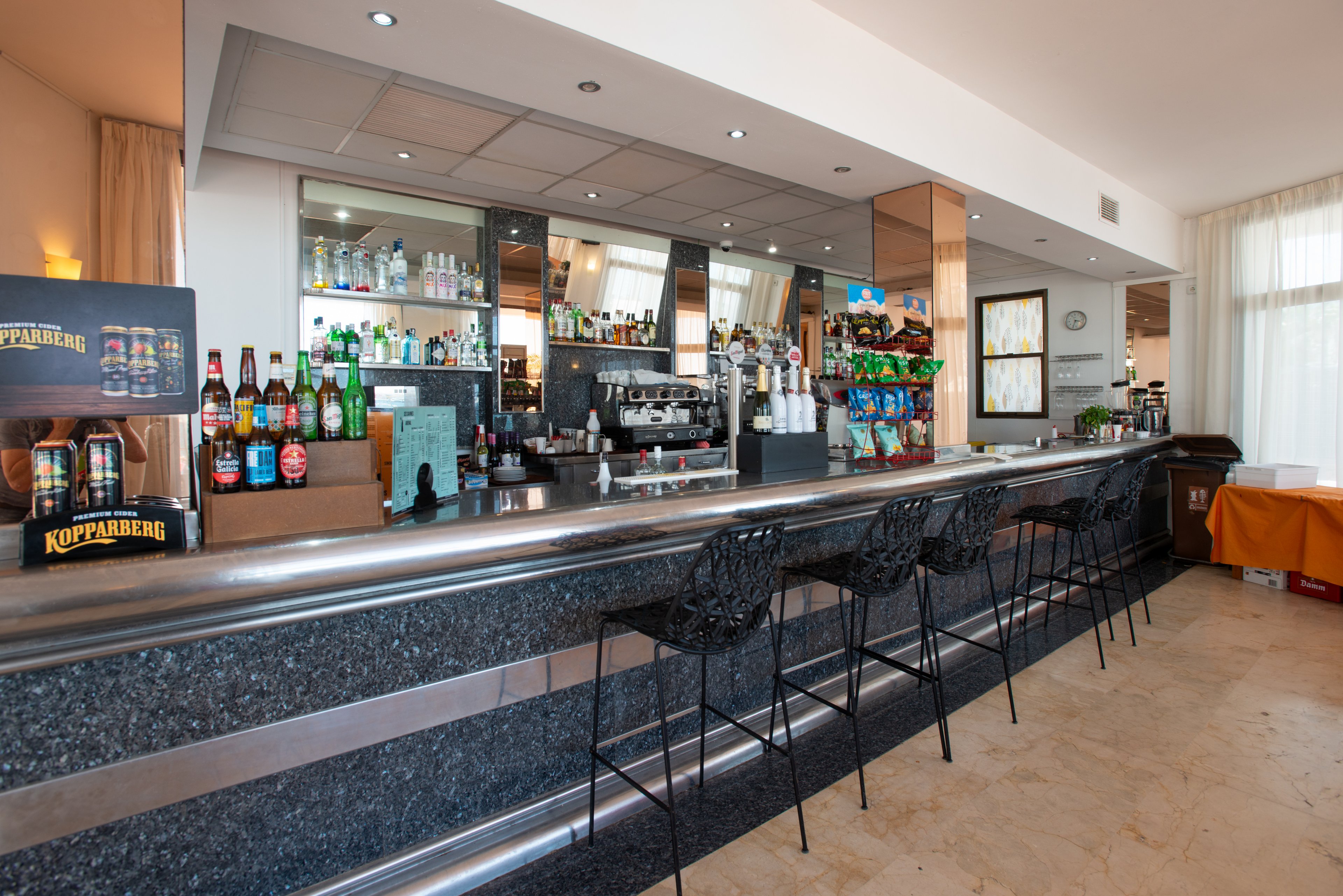 A contemporary bar counter is lined with several empty black stools in a bright, open interior. The back bar shelves display a wide selection of spirits, liqueurs, and bottled drinks, with a coffee machine and bar equipment at the center. Colorful snack displays and drink bottles add visual interest to the otherwise neutral, polished surfaces. Large windows and light walls create an airy, welcoming atmosphere suitable for hotel guests or casual visitors.
