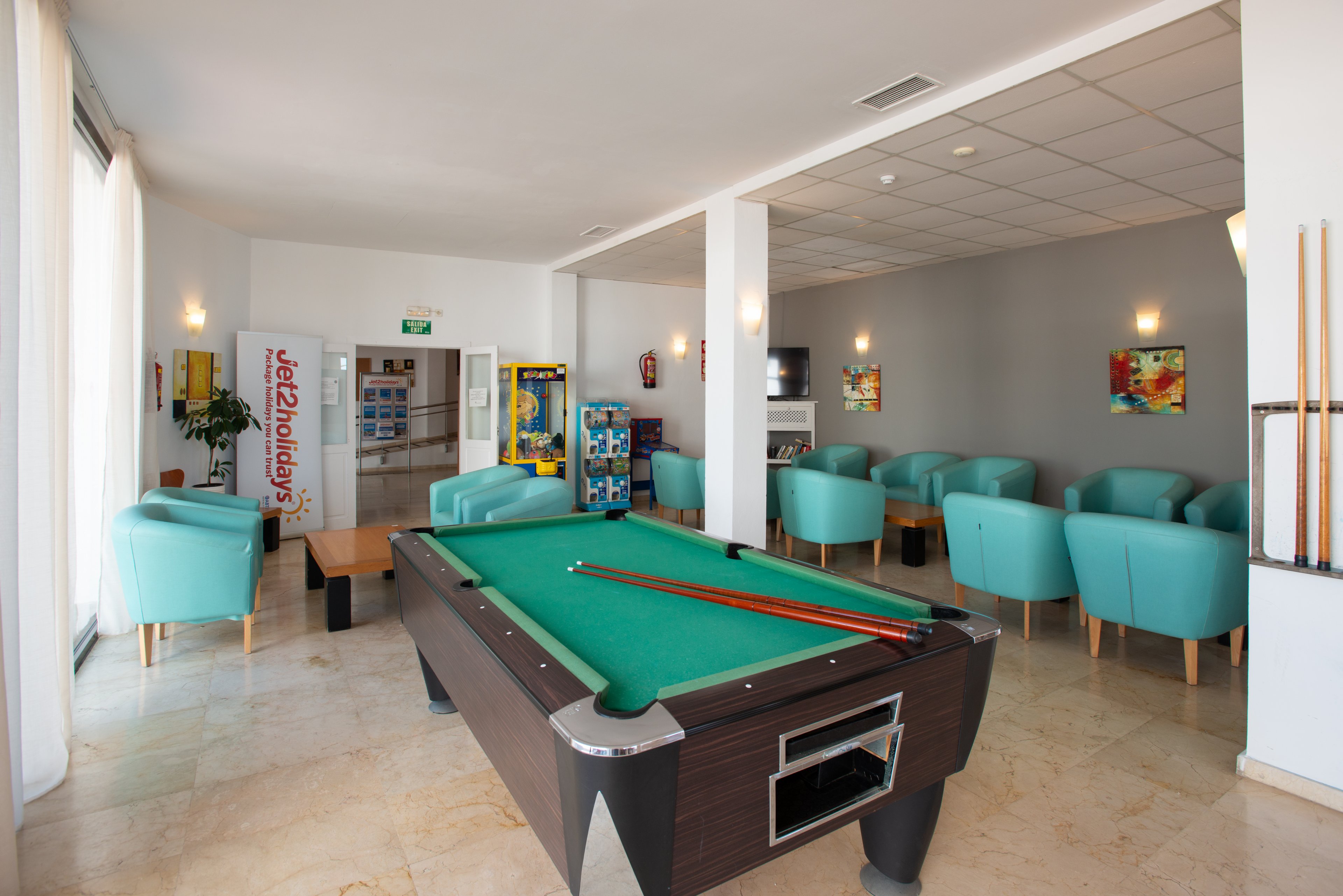 A modern hotel lounge features a central pool table with cues laid neatly on the green felt. Turquoise armchairs are arranged in cozy seating areas around small tables, creating a relaxed social space. Large windows on the left bring in natural light, while wall sconces and colorful artwork add warmth to the room. In the background, vending machines and a doorway suggest additional guest amenities.