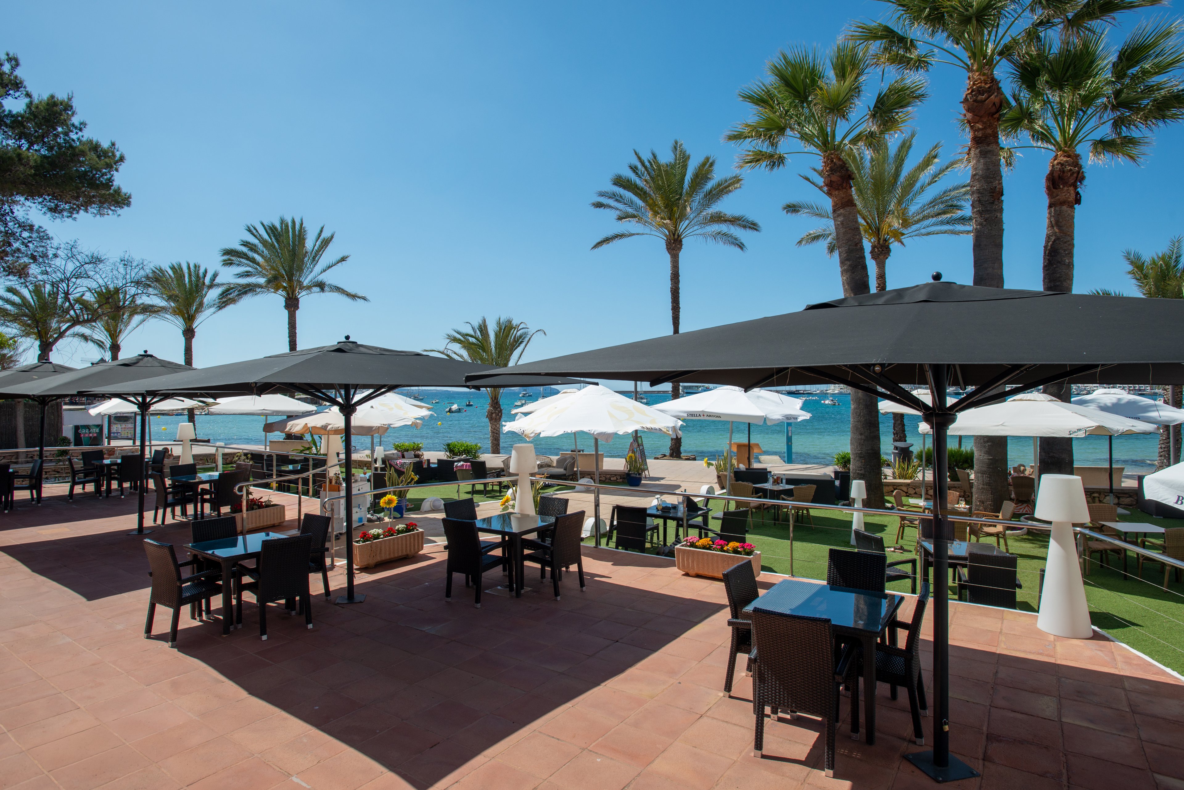 An open-air restaurant terrace overlooks a calm seaside with clear blue skies. Black parasols shade dark tables and chairs arranged on a terracotta-tiled floor. White umbrellas and modern outdoor lamps line the edge of the patio near a green lawn. Tall palm trees frame the scene, creating a relaxed Mediterranean resort atmosphere.