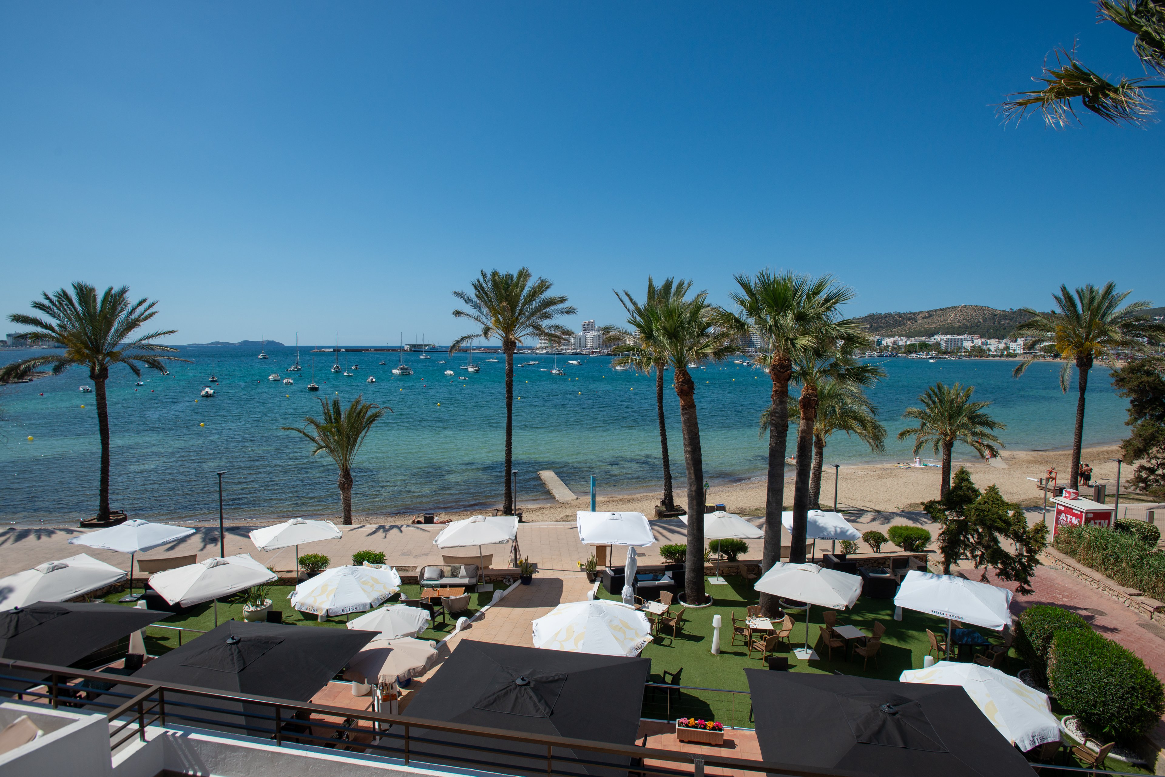 A coastal resort terrace overlooks a sandy beach lined with tall palm trees. White and black sun umbrellas shade outdoor seating areas on a manicured lawn. Beyond the beach, calm blue water is dotted with small boats and yachts under a clear sky. Low hills and distant buildings frame the bay, creating a relaxed Mediterranean holiday atmosphere.