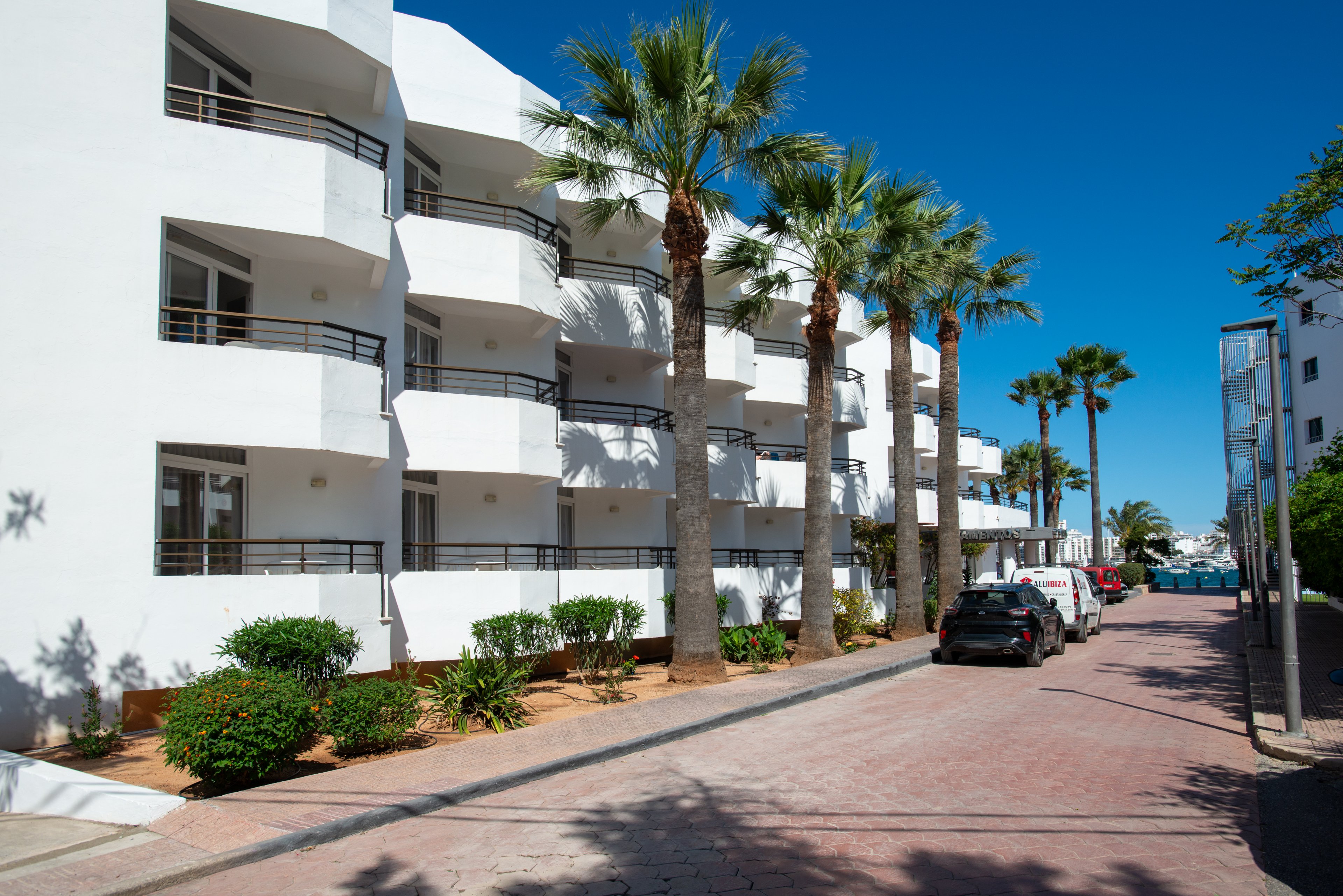 A row of modern white apartment buildings lines a quiet street under a clear blue sky. Tall palm trees and neatly trimmed shrubs decorate the sidewalk in front of the residences. Parked cars are visible along the paved road, suggesting a calm residential or holiday neighborhood. The bright sunlight and clean architecture create a warm, Mediterranean-style atmosphere.