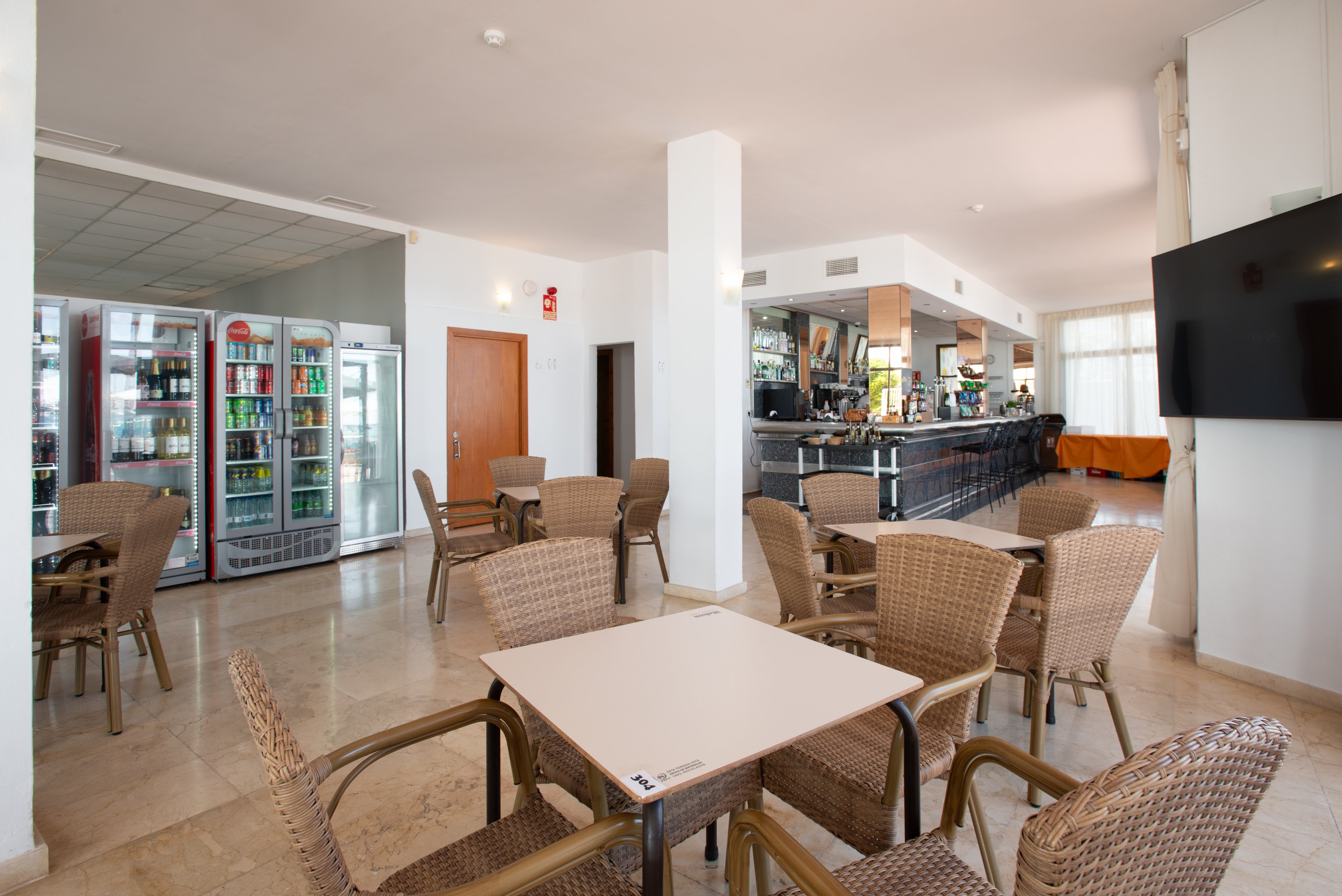 Spacious bar interior featuring several wicker tables and chairs arranged on a polished floor. A long counter with bar stools and bottles in the background suggests a casual dining or drink service area. Large glass-front refrigerators display assorted beverages along one wall. Natural light enters through wide windows, creating an airy and welcoming atmosphere suitable for guests to relax.
