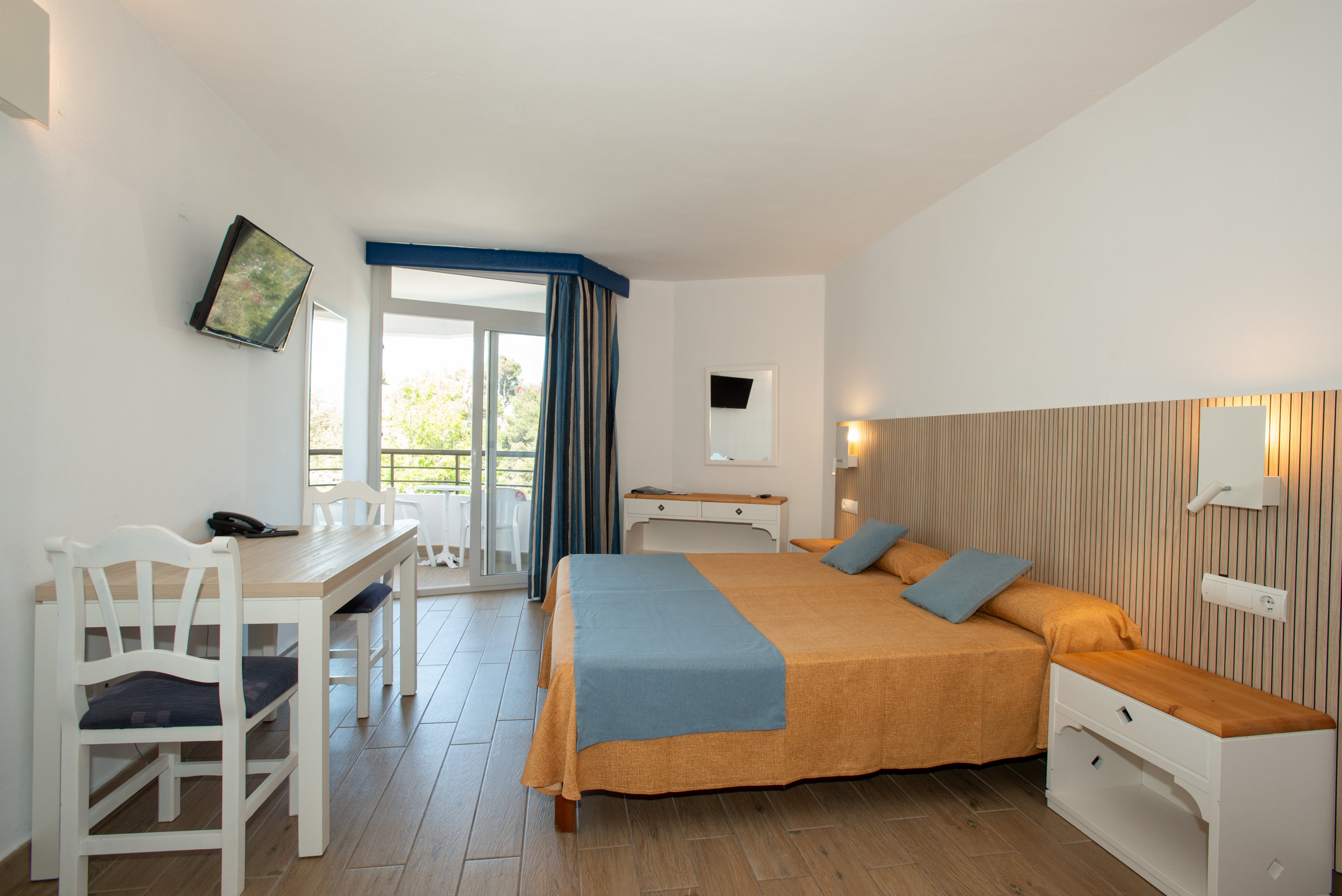 A tidy hotel room features a large double bed with an orange bedspread and blue accent pillows. The space includes a white desk with a chair, a wall-mounted television, and a small dresser with a mirror. Floor-to-ceiling glass doors open onto a balcony with outdoor seating and a view of greenery. Light wood flooring and white walls create an airy, modern atmosphere with blue curtains adding contrast.