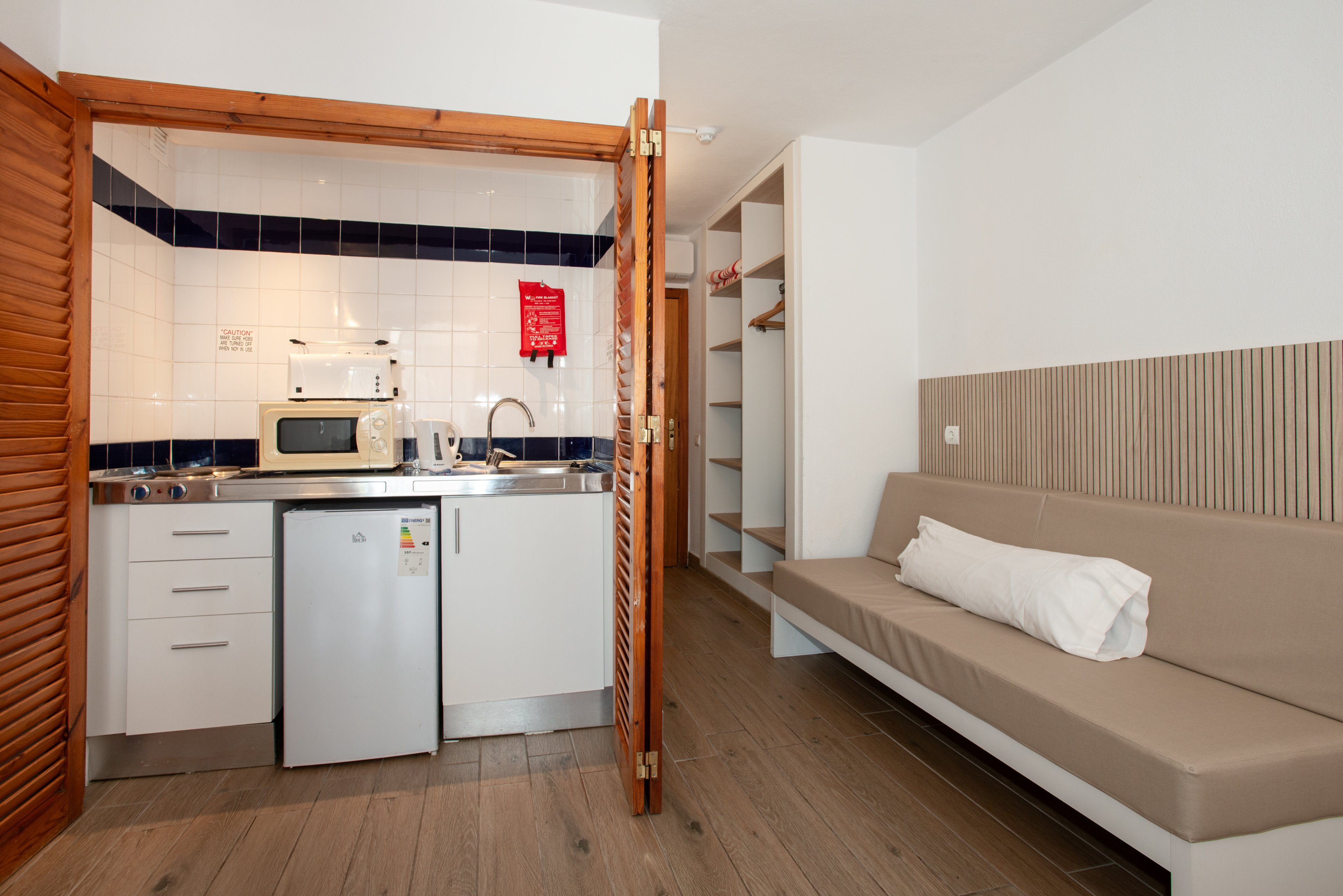 A compact studio kitchenette features white cabinets, a small refrigerator, and a microwave set against a tiled backsplash. Wooden louvered doors frame the kitchen area, which opens into a simple living space. A beige bench-style sofa with a rolled white towel sits opposite an open closet with shelves and hanging space. The room has light wood flooring and neutral walls, creating a clean and functional atmosphere.