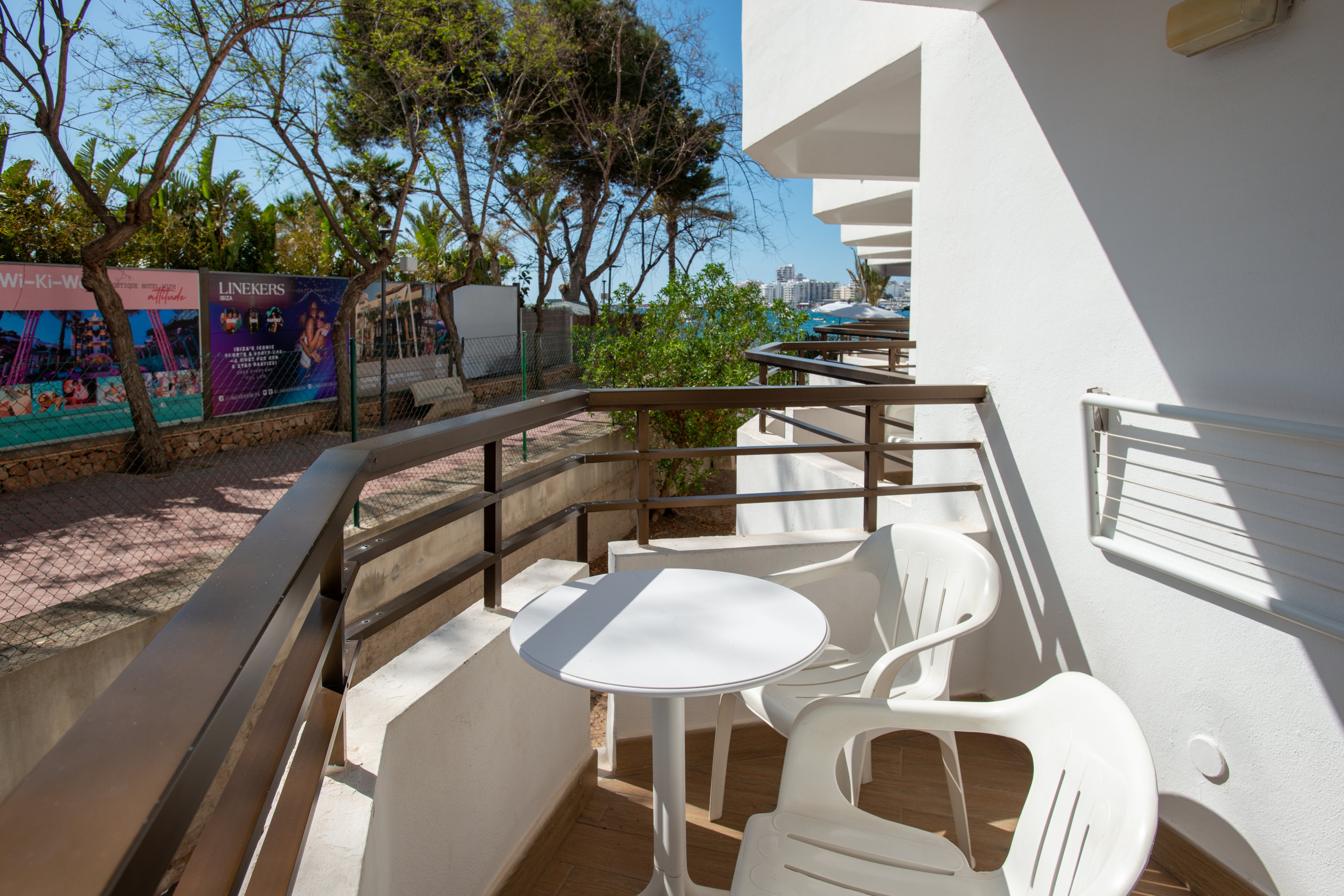 A small balcony features a round white table and two white plastic chairs in bright sunlight. The balcony is attached to a modern white building with metal railings. In the background, a pedestrian walkway, trees, and colorful street posters are visible. The scene suggests a warm, relaxed holiday setting near an urban or coastal area.