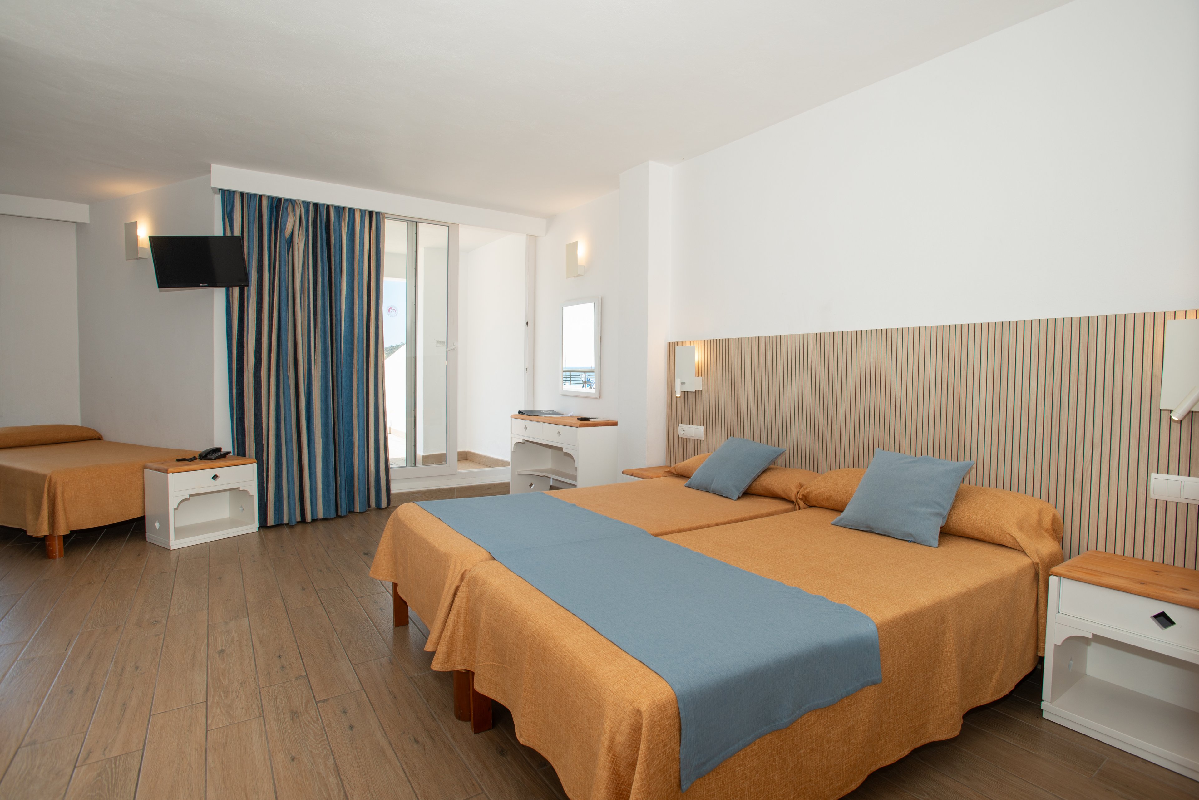 Spacious hotel room featuring a large double bed in the foreground and a single bed in the background. The beds are dressed in warm tan bedspreads with light blue runners and cushions. A wall-mounted television, bedside tables, and modern wall lights add comfort and functionality. Floor-to-ceiling striped curtains lead to a balcony or window area, bringing natural light into the clean, minimalist interior.
