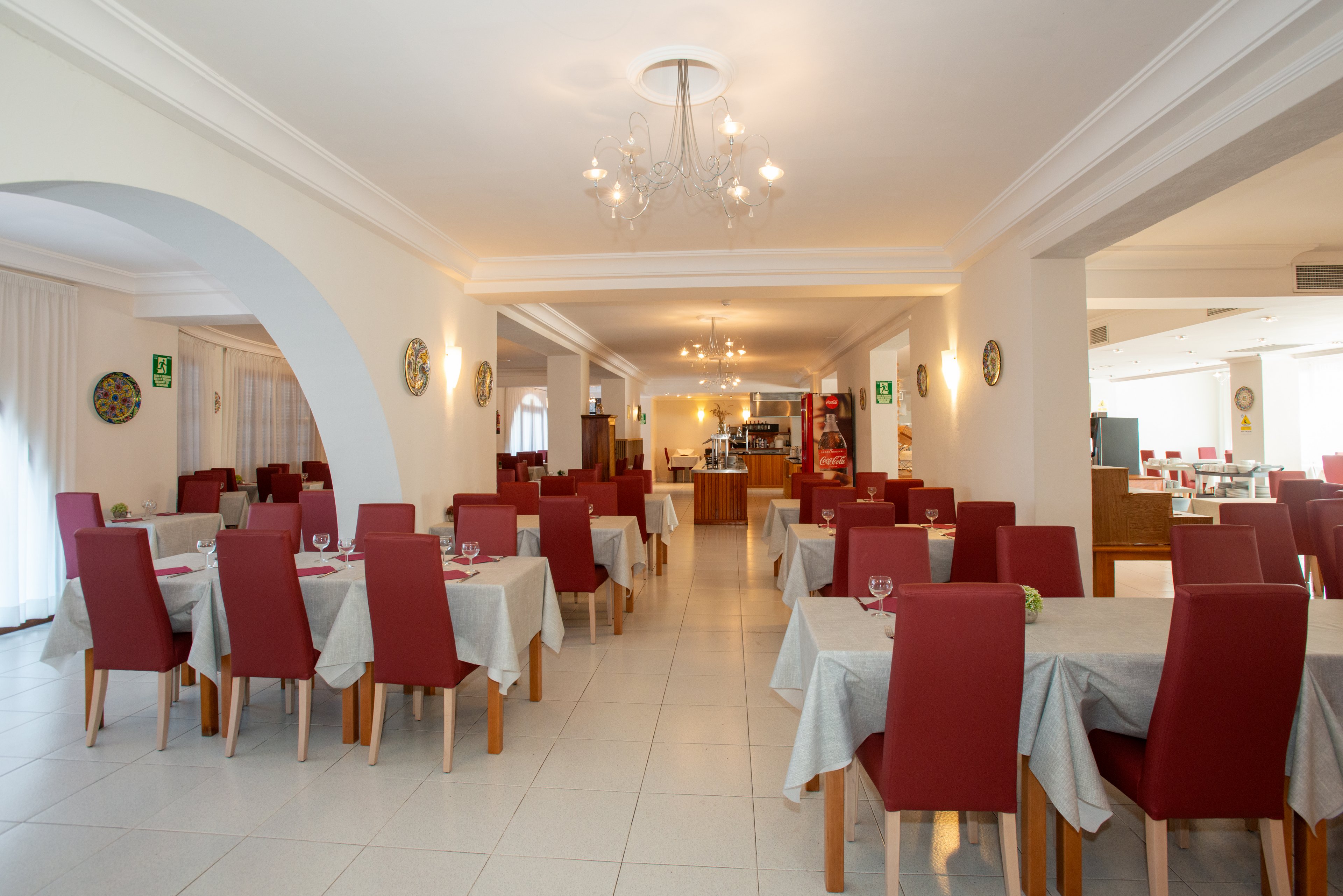 Spacious restaurant dining room with neatly arranged tables and red upholstered chairs. White tablecloths and place settings with glasses create a formal yet welcoming atmosphere. Large windows and light-colored walls brighten the space, while chandeliers add an elegant touch. The room appears ready for guests, with a buffet or service area visible in the background.