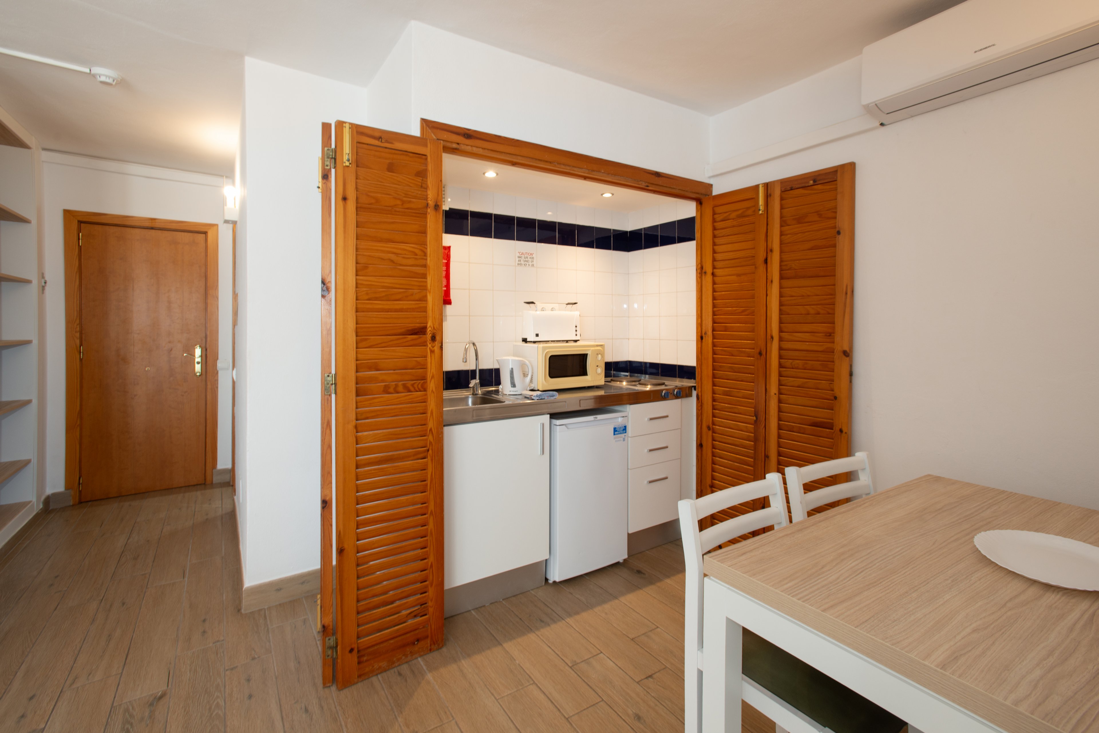 A small white kitchen is partially enclosed by wooden folding doors in a compact studio apartment. The kitchenette includes a microwave, kettle, sink, and a mini refrigerator beneath a tiled backsplash. A simple dining table with chairs sits in the foreground on light wood flooring. Built-in shelves and a closed wooden door are visible in the background, creating a clean and functional interior space.