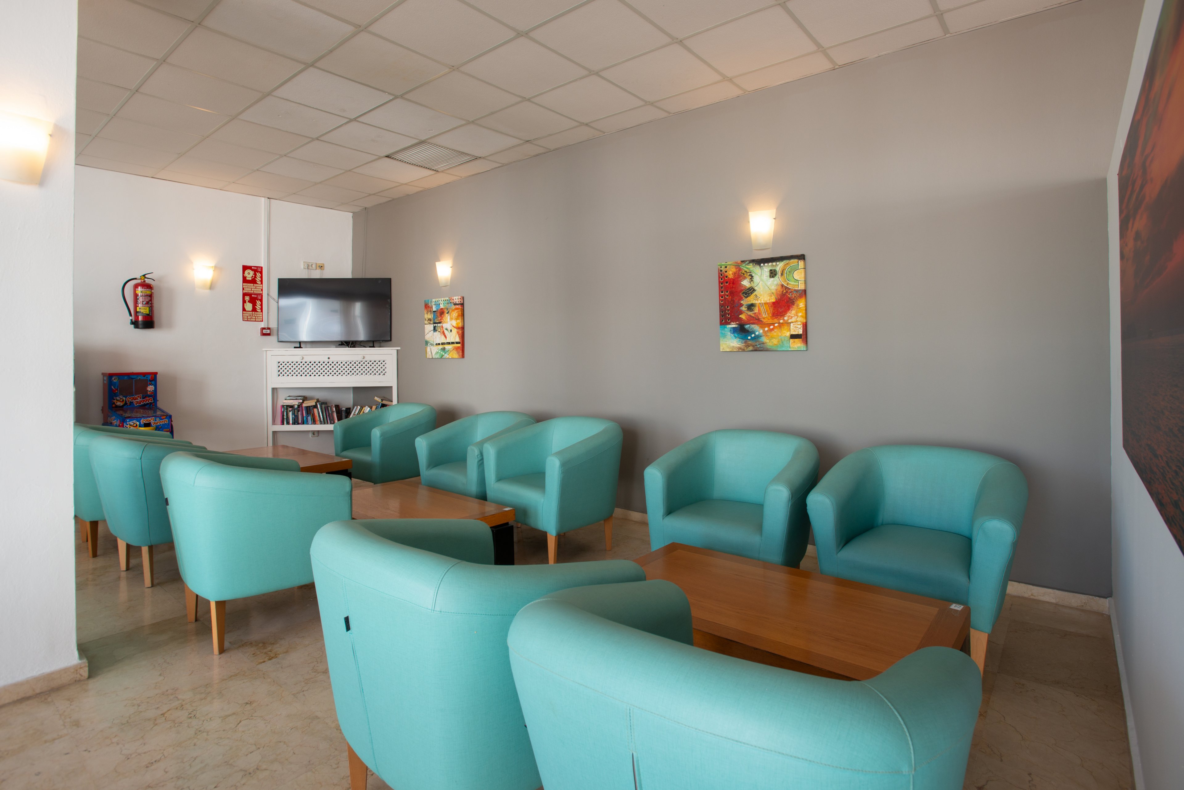 A small lounge area features several turquoise armchairs arranged around light wooden coffee tables. The space has neutral walls, a tiled ceiling, and wall-mounted lights creating a soft, welcoming glow. A television and bookshelf are visible against the far wall, along with colorful abstract artwork. Fire safety equipment and signage are mounted near the entrance, indicating a public or hotel setting.