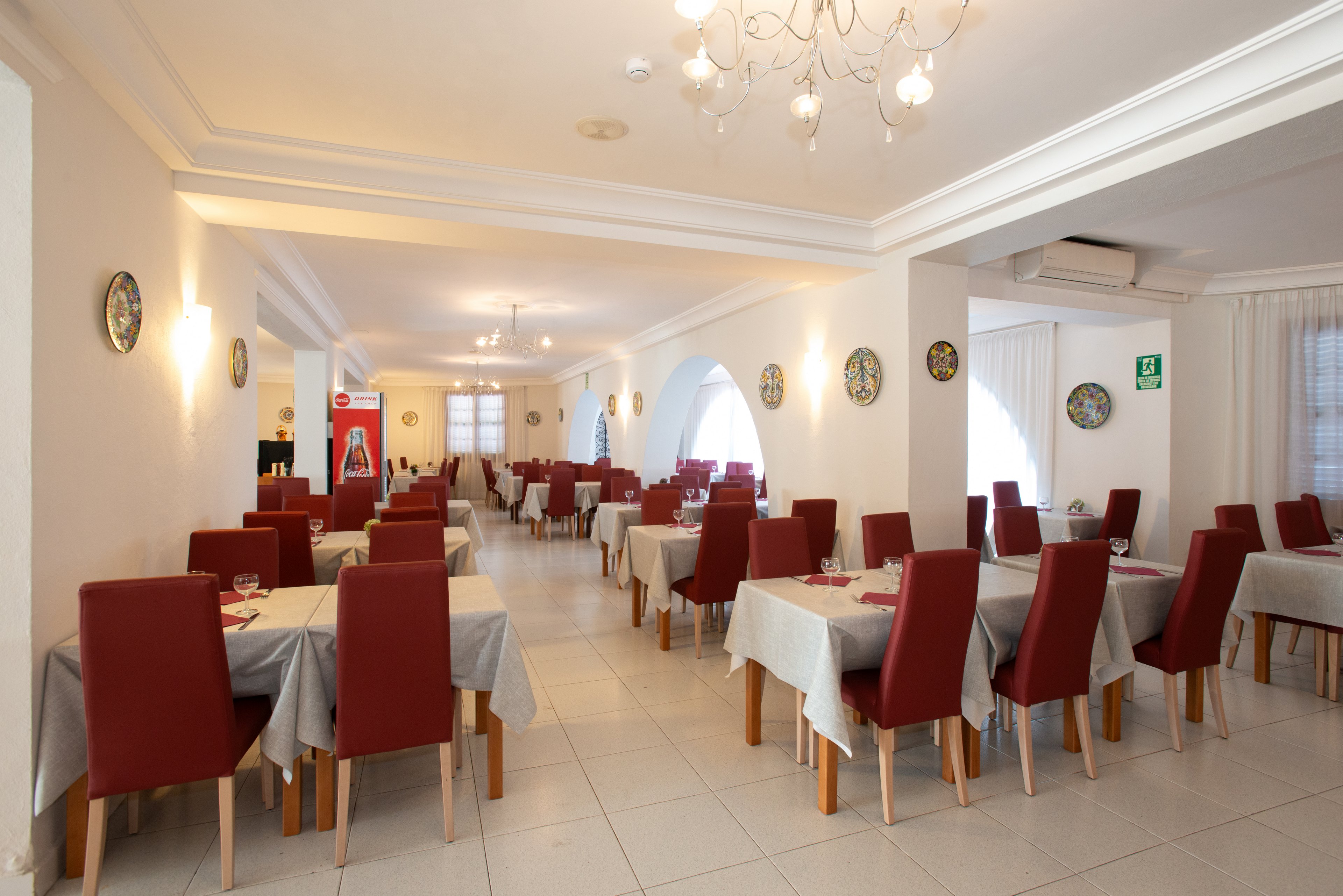 Spacious restaurant dining room with neatly arranged tables and red upholstered chairs. White walls, tiled floor, and large archways create an airy, minimalist atmosphere. Tables are covered with light gray tablecloths and set with glasses and napkins, but no diners are present. Warm ceiling lights and wall sconces illuminate the space, highlighting decorative plates on the walls and a calm, orderly setting.