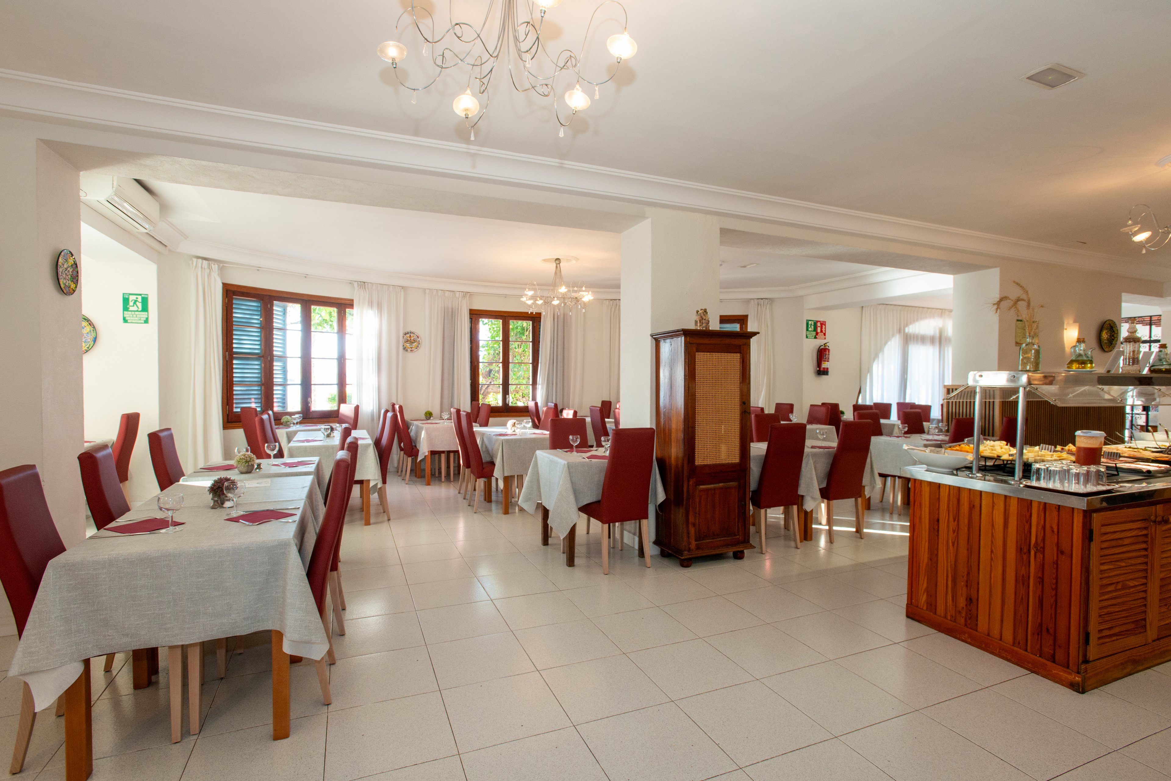 Spacious restaurant dining room with neatly arranged tables and red upholstered chairs. White tablecloths and tiled floors create a clean, minimalist look. Large windows with wooden frames let in natural light and offer a view of greenery outside. A wooden buffet station with covered dishes stands to the right, suggesting a self-service or breakfast area. Chandeliers and soft neutral colors contribute to a calm, welcoming atmosphere.