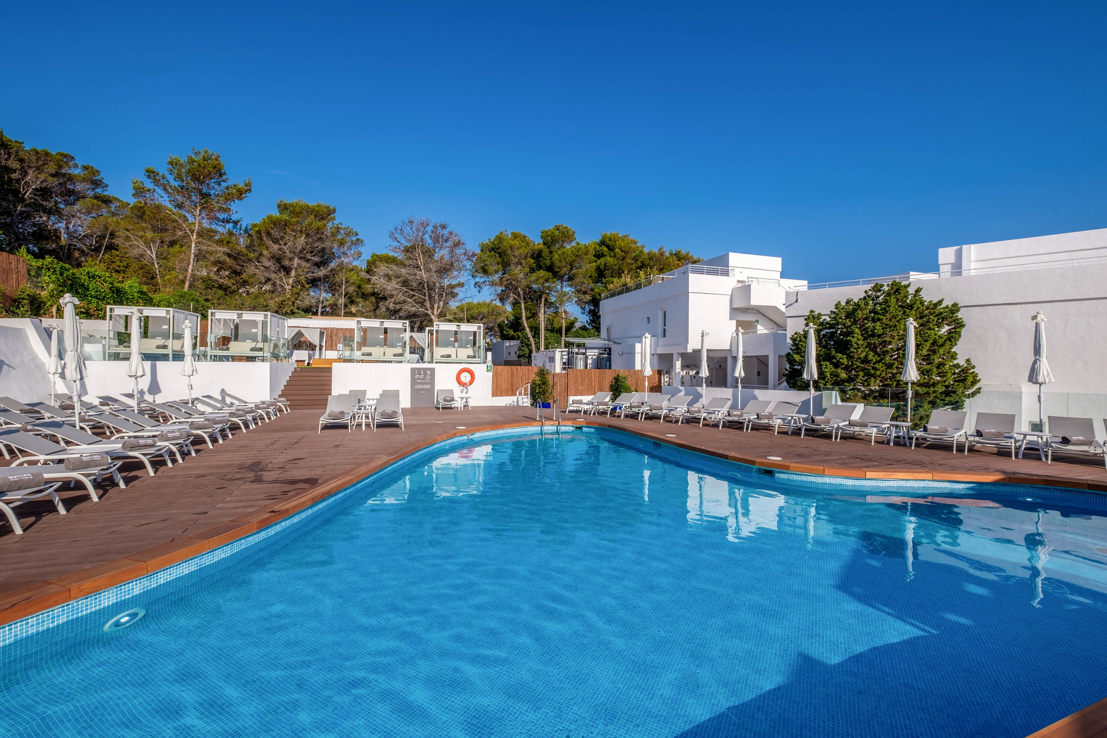 A clear blue swimming pool is surrounded by numerous white sun loungers and umbrellas at a modern resort. White buildings with clean lines and glass railings frame the pool area, set against a bright blue sky. Trees and greenery rise behind the structures, adding a natural backdrop to the scene. The overall atmosphere suggests a calm, sunny day ideal for relaxation and leisure by the water.