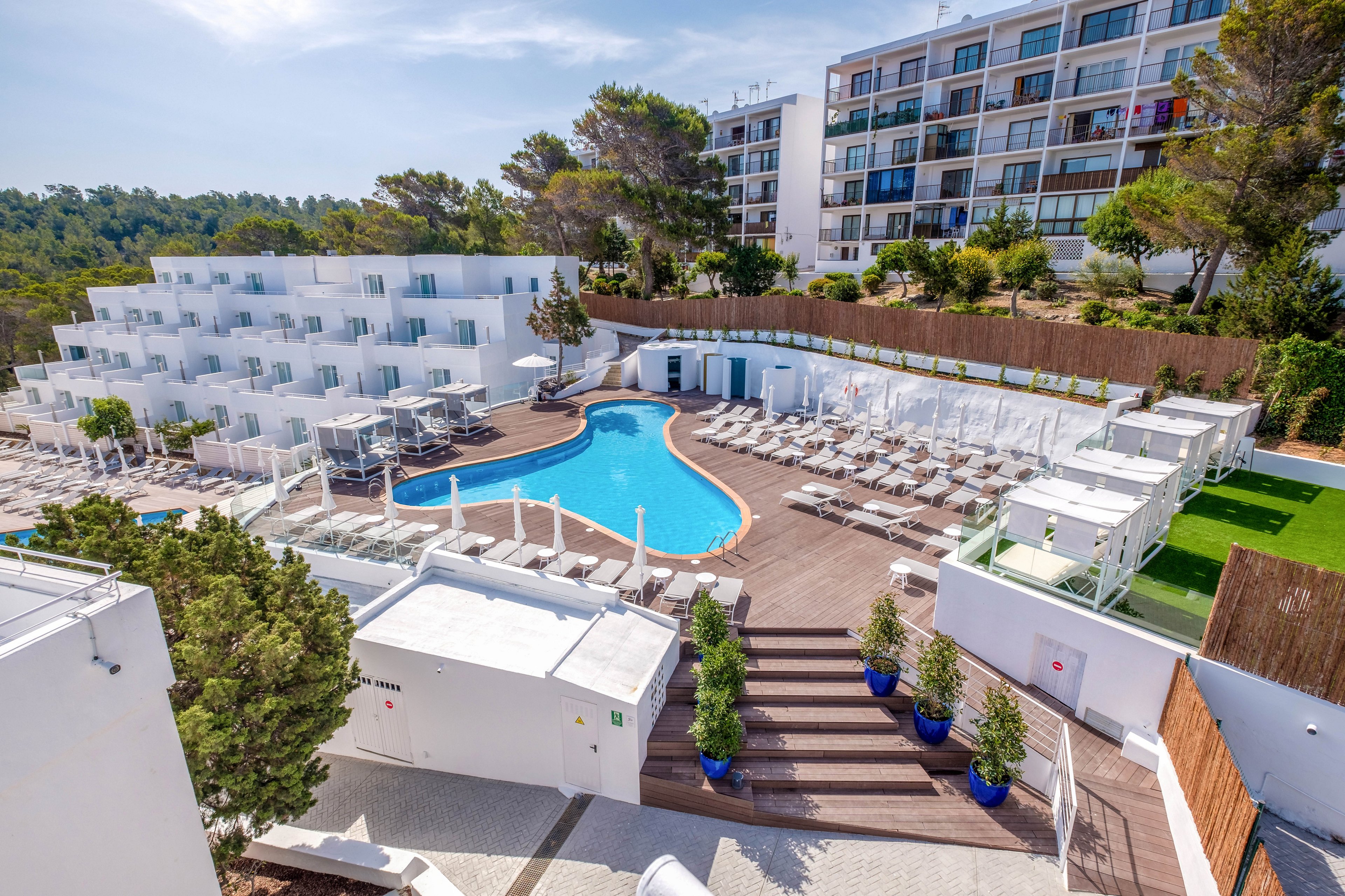 A contemporary resort features a central outdoor swimming pool surrounded by numerous white sun loungers and umbrellas. Tiered white buildings and a taller modern apartment-style structure frame the pool area. Green trees and landscaped plants add contrast to the bright wooden deck and white architecture. The scene is captured from an elevated viewpoint on a sunny day, emphasizing the clean, minimalist design and vacation atmosphere.